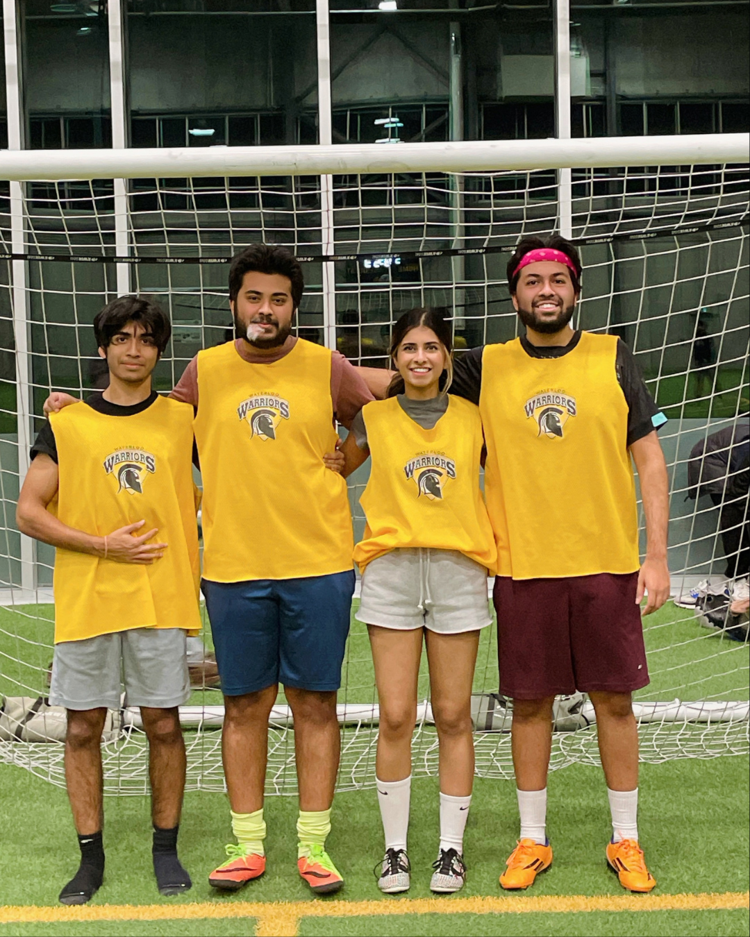 Four people wearing yellow Waterloo Warriors pinnies pose together in front of an indoor soccer goal on artificial turf.