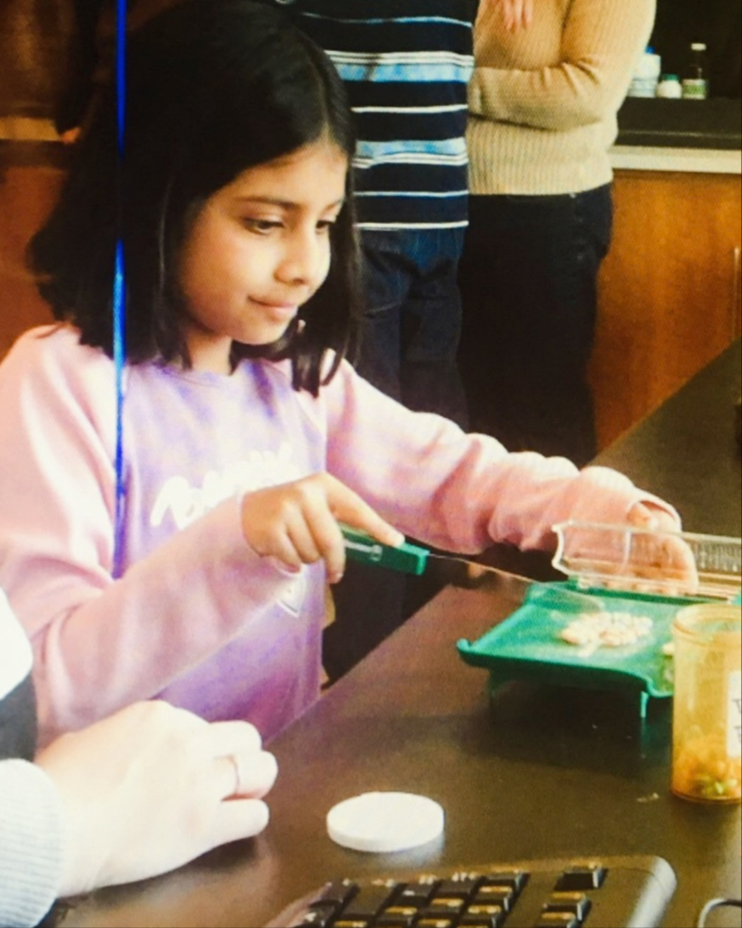 A child in a pink sweatshirt uses a knife to cut food on a small green cutting board at a table during an indoor activity, with several people standing nearby.