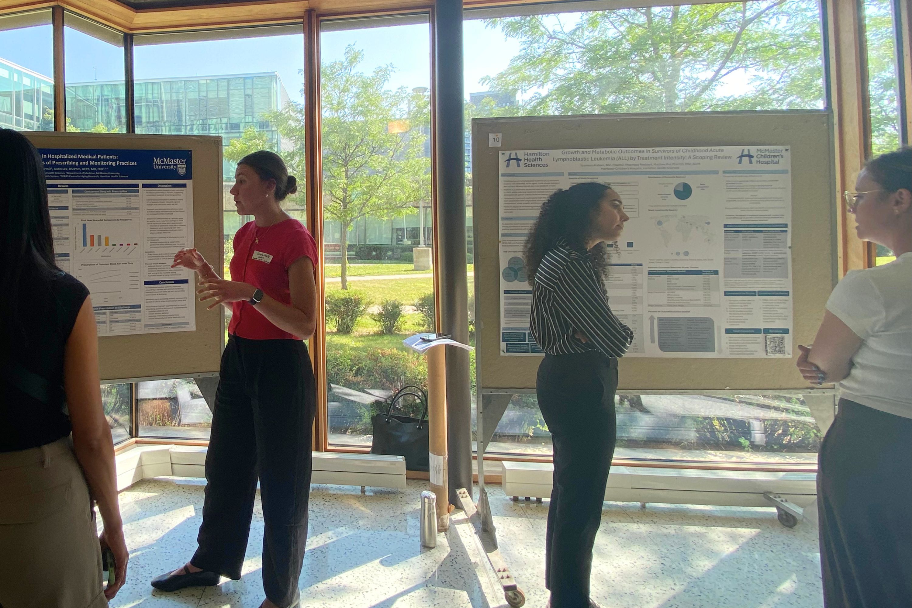Several participants stand in front of research posters displayed on presentation boards, discussing findings during a poster session. The event takes place in a sunlit hallway with floor-to-ceiling windows overlooking trees and campus buildings in the background.