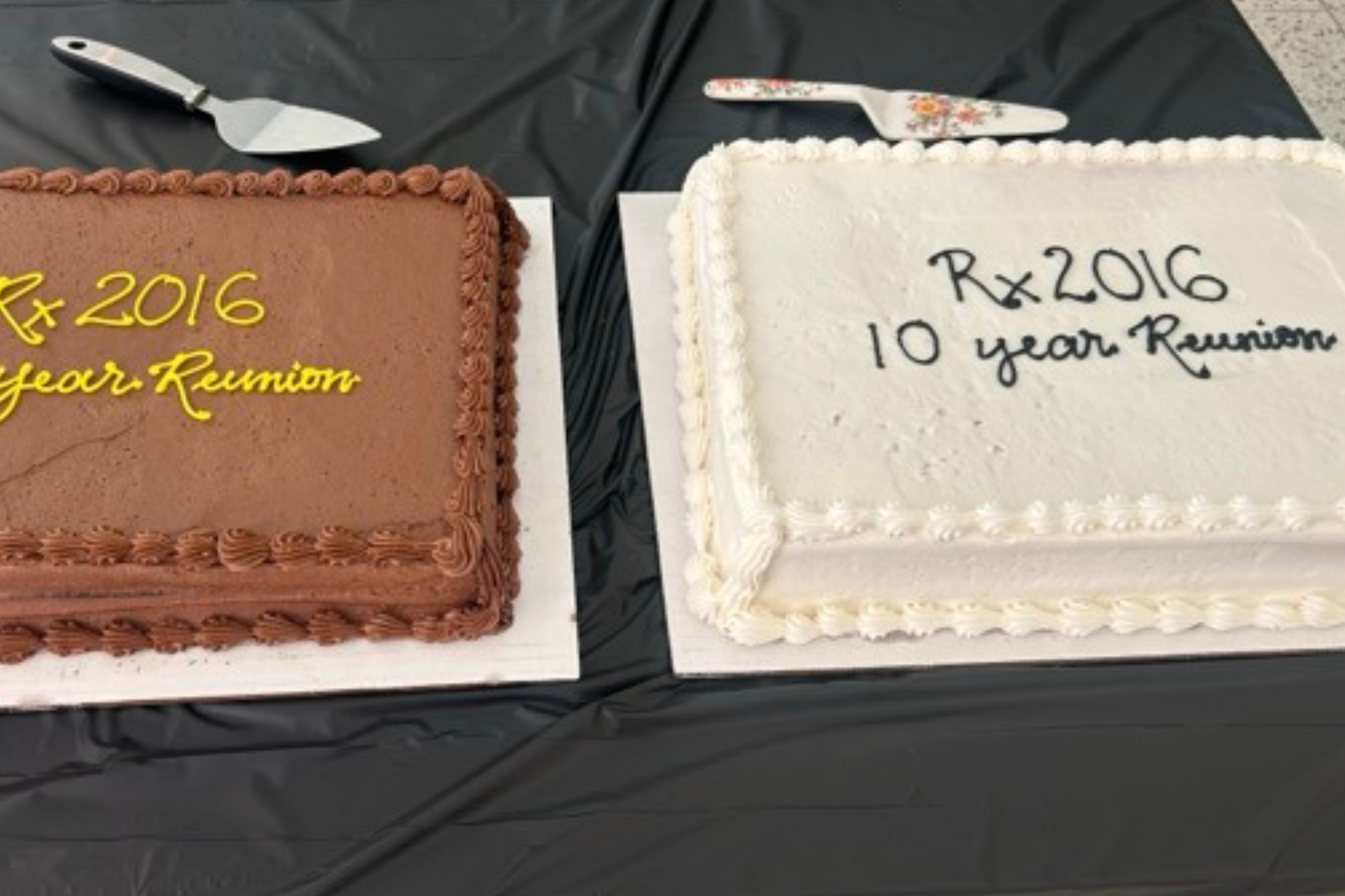Two rectangular cakes, one chocolate and one white, sit side by side on a black tablecloth. Both are decorated with “Rx 2016 10 year Reunion,” with cake servers resting above them.