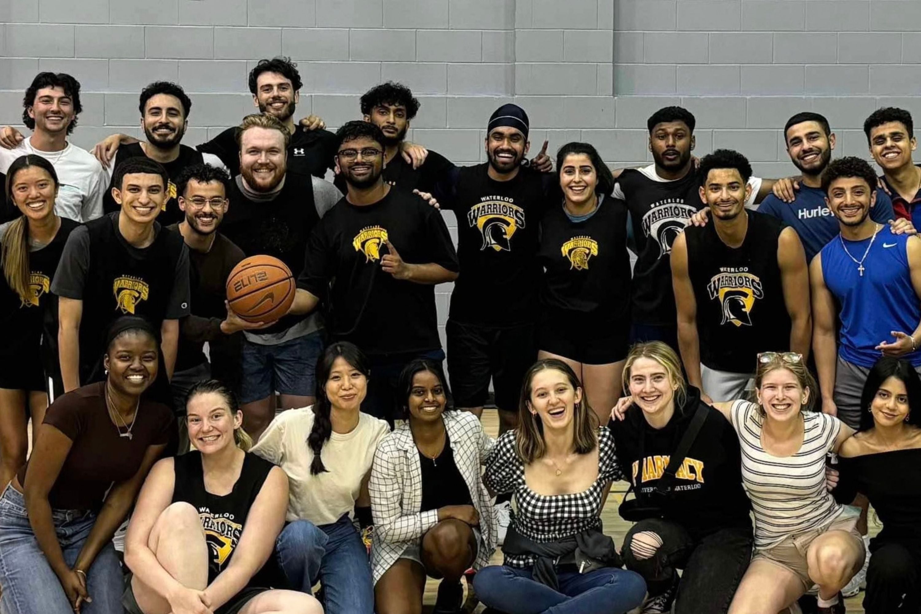 A large group poses together on a basketball court, with several people wearing Waterloo Warriors shirts and one person holding a basketball.