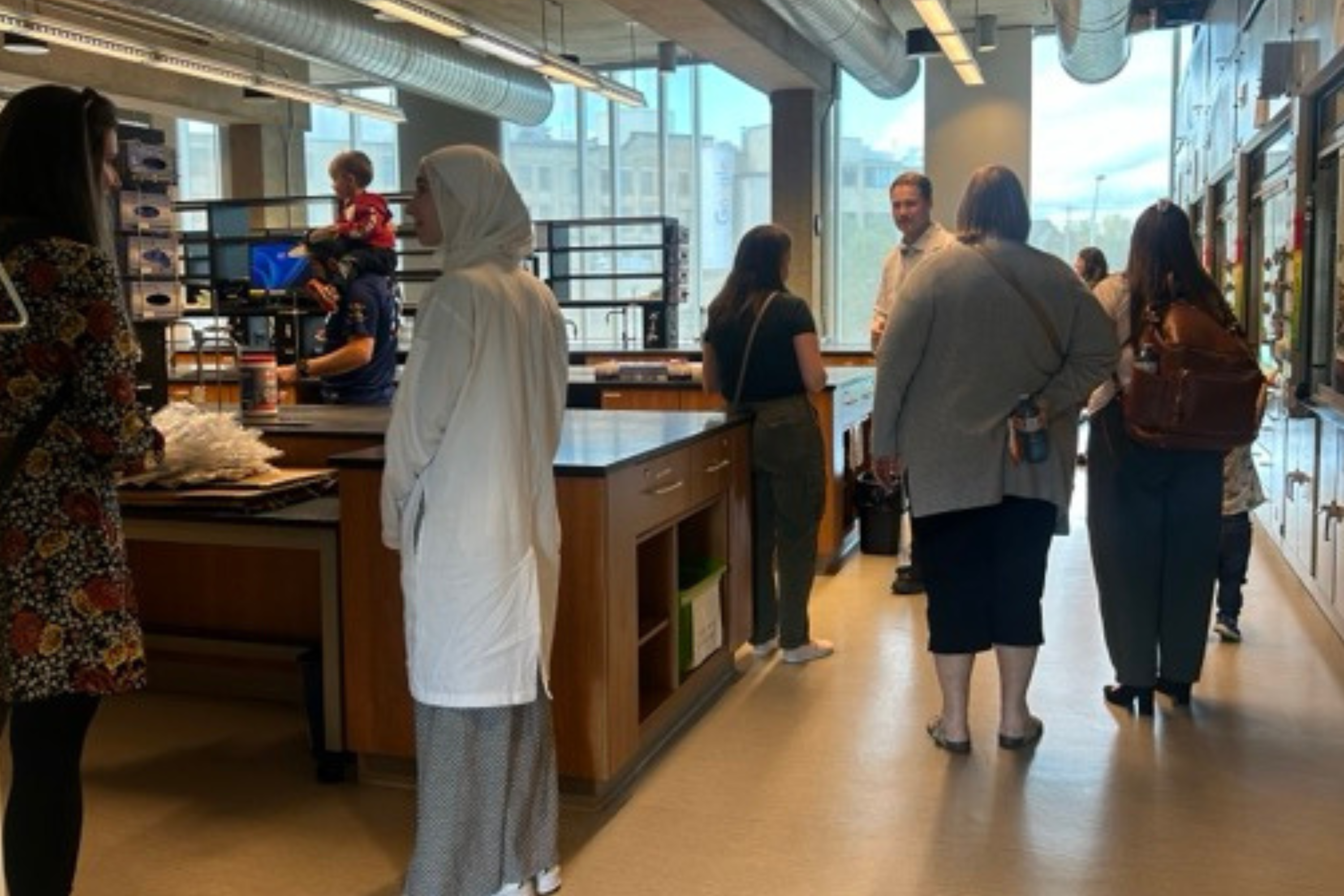 A group of people gather inside a bright teaching laboratory with long workbenches, cabinets and lab equipment, while several people talk near the centre and others stand around the room.