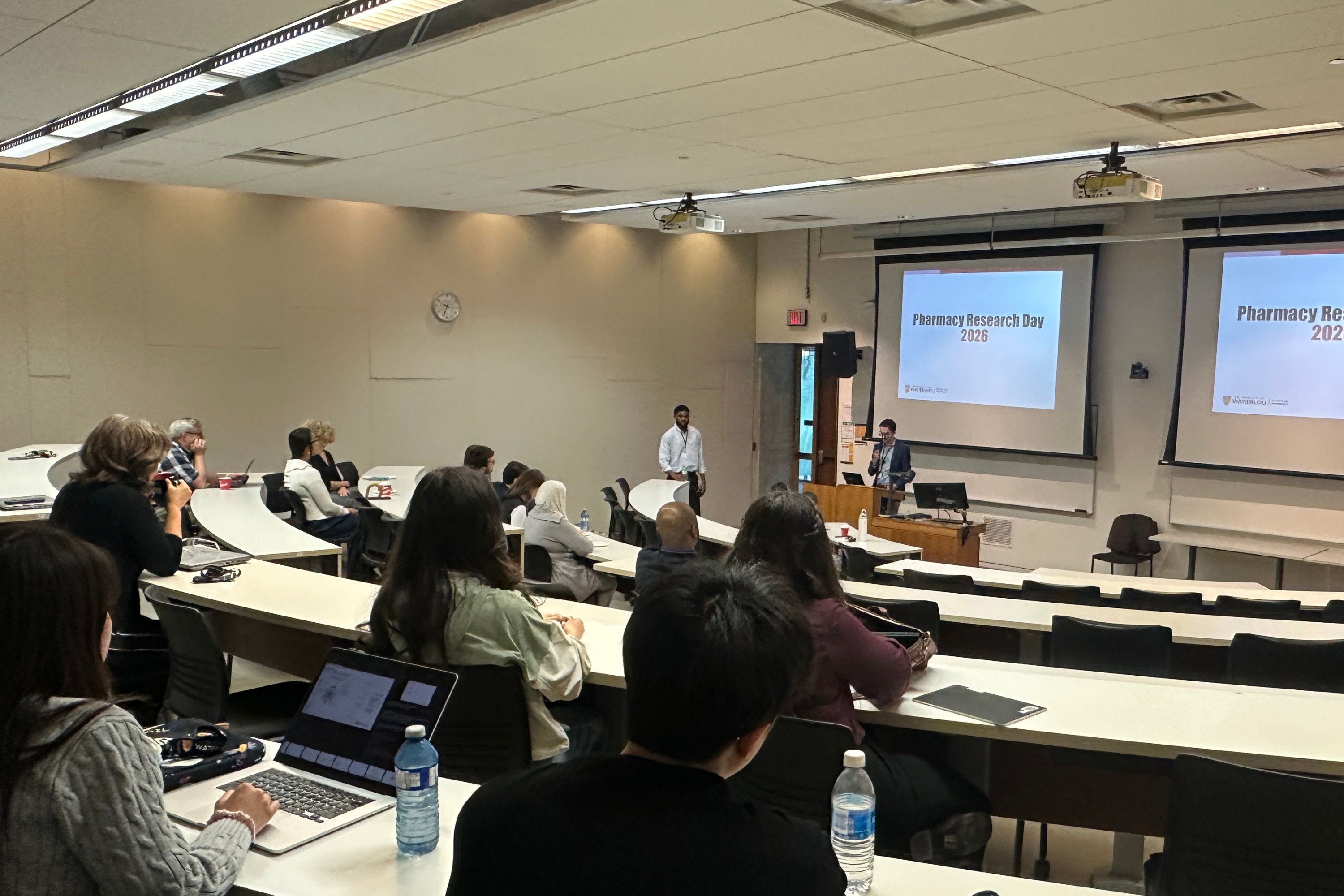 Tiered lecture hall with attendees seated at long desks using laptops and notebooks, facing a speaker at a podium. A presentation slide on screen reads “Pharmacy Research Day 2026.”
