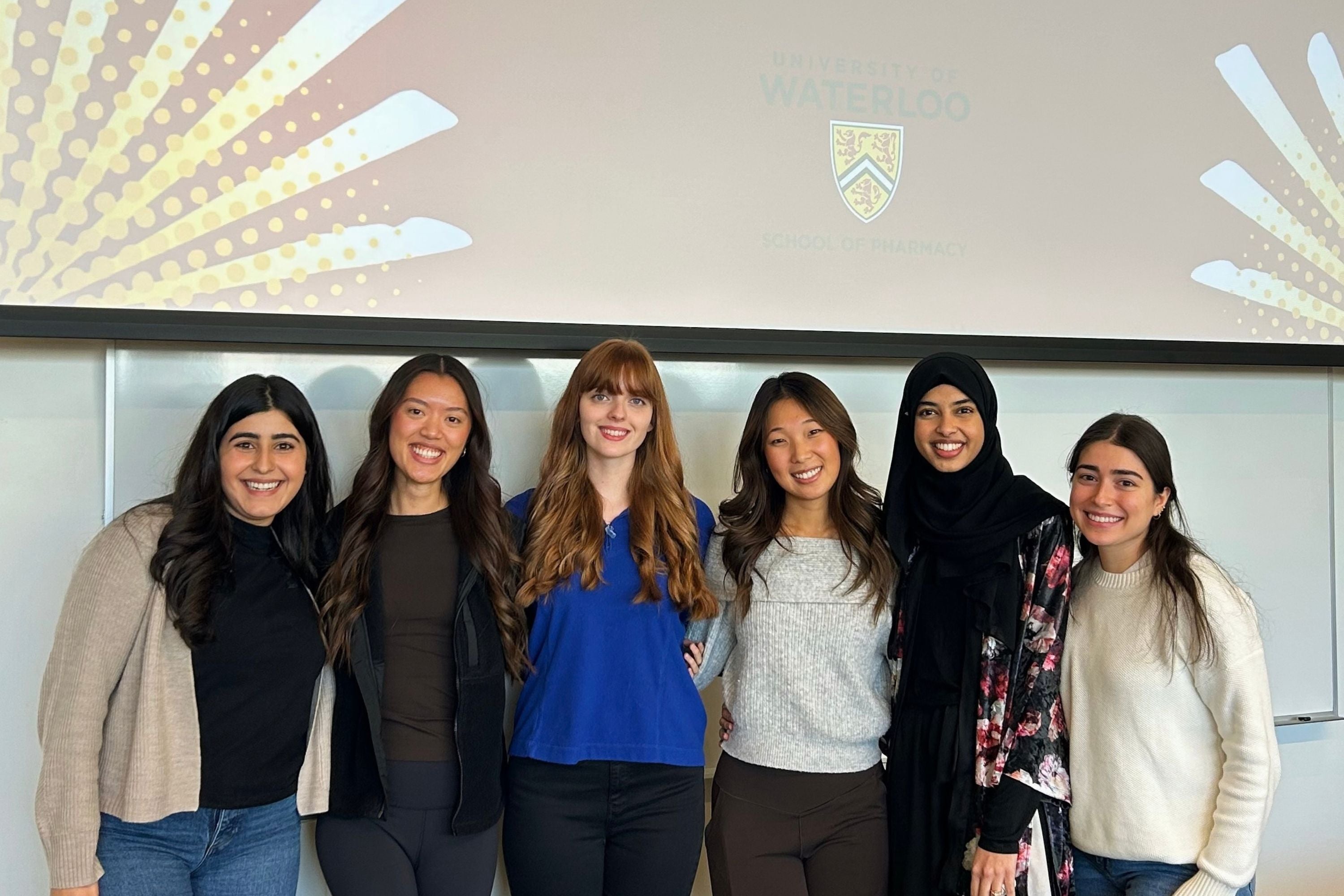 Six people pose together in a classroom in front of a projection screen displaying the University of Waterloo School of Pharmacy logo