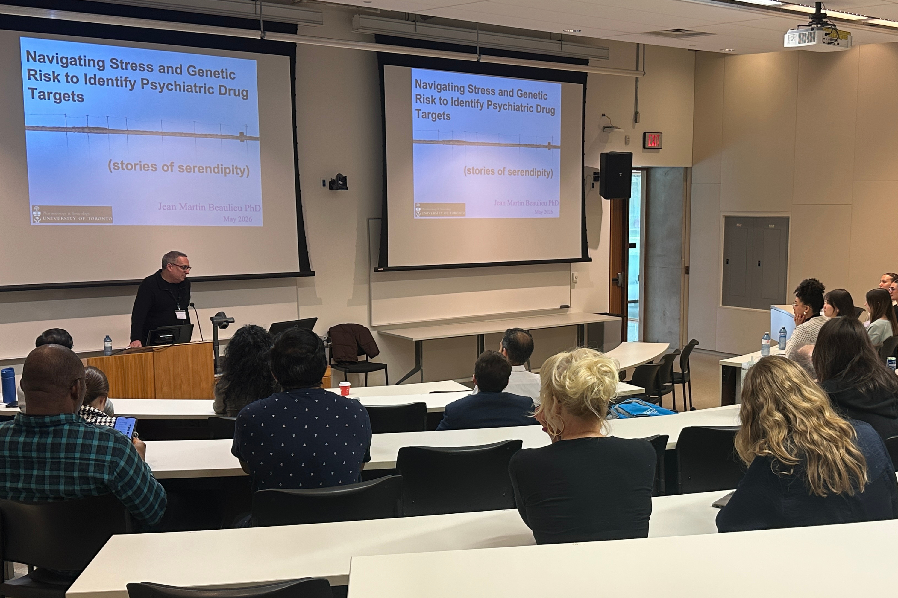 A speaker presents at the front of a lecture hall beside two large projection screens displaying a slide titled “Navigating Stress and Genetic Risk to Identify Psychiatric Drug Targets,” while attendees sit and listen.