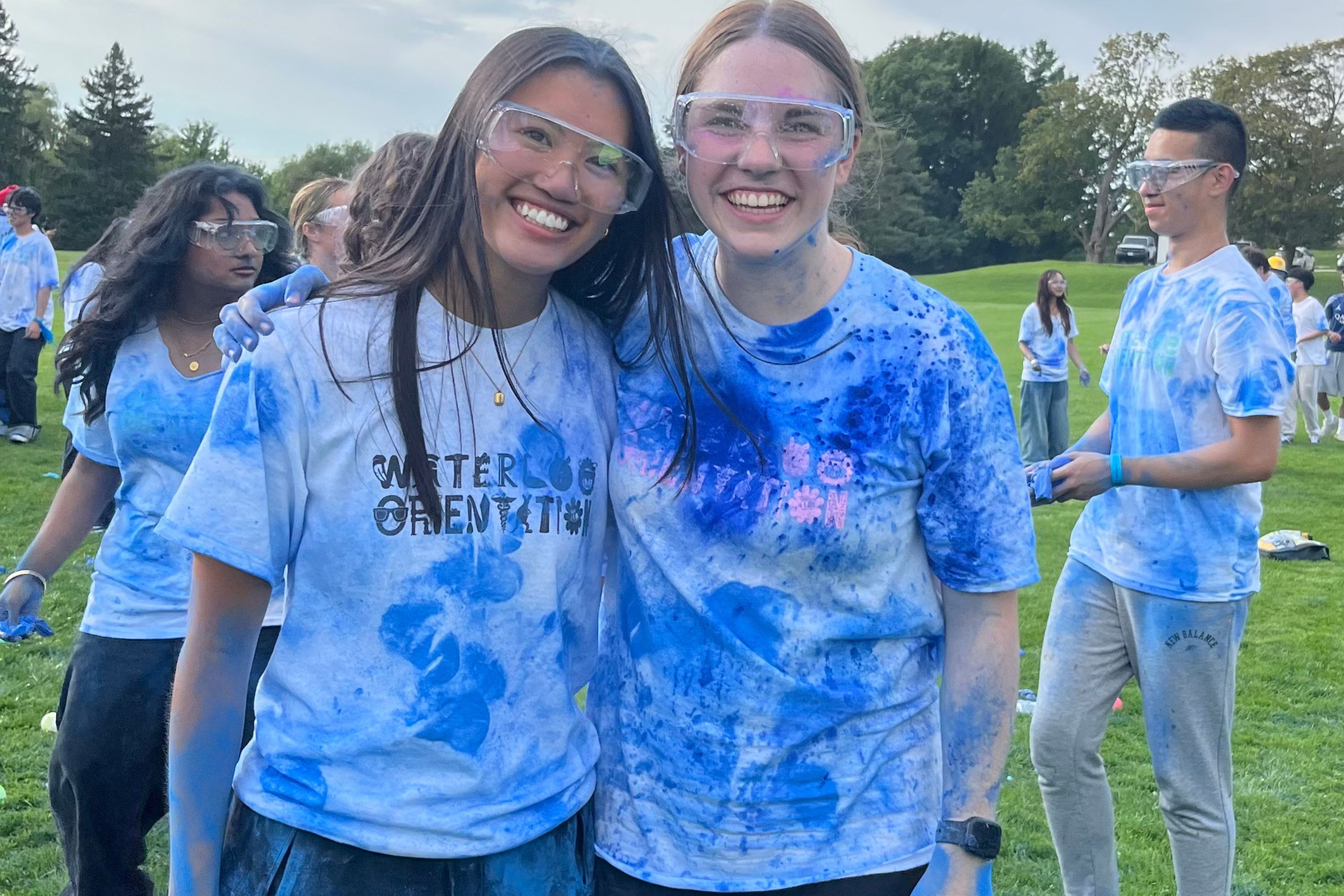 Two people wearing safety goggles and blue-stained shirts pose together outdoors, with other participants gathered on a grassy field in the background