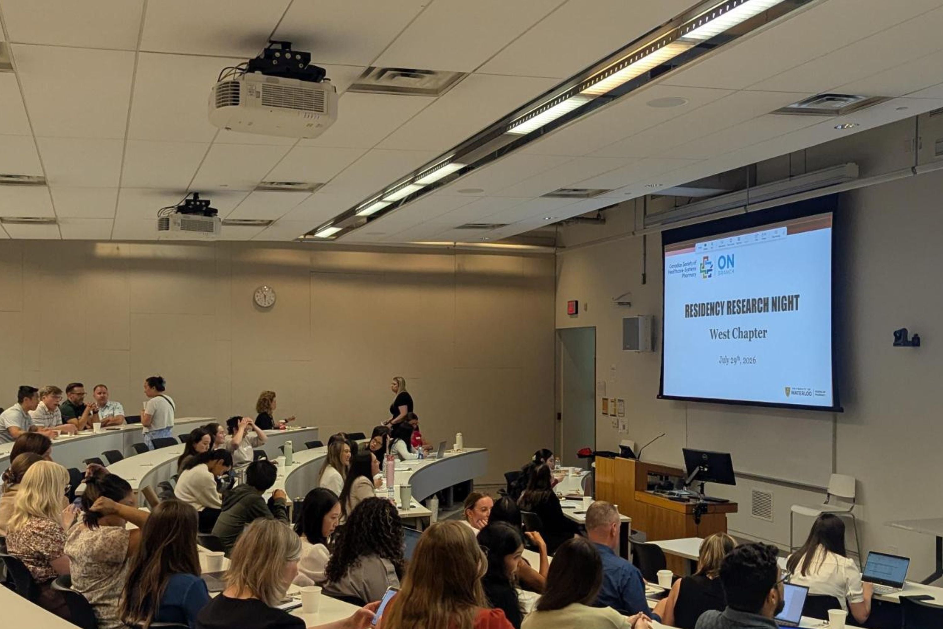 Participants sit and converse in a tiered lecture hall before a research event. A large projection screen at the front of the room displays a title slide reading “Residency Research Night, West Chapter, July 29, 2026,” alongside organizational logos and a podium for presenters.