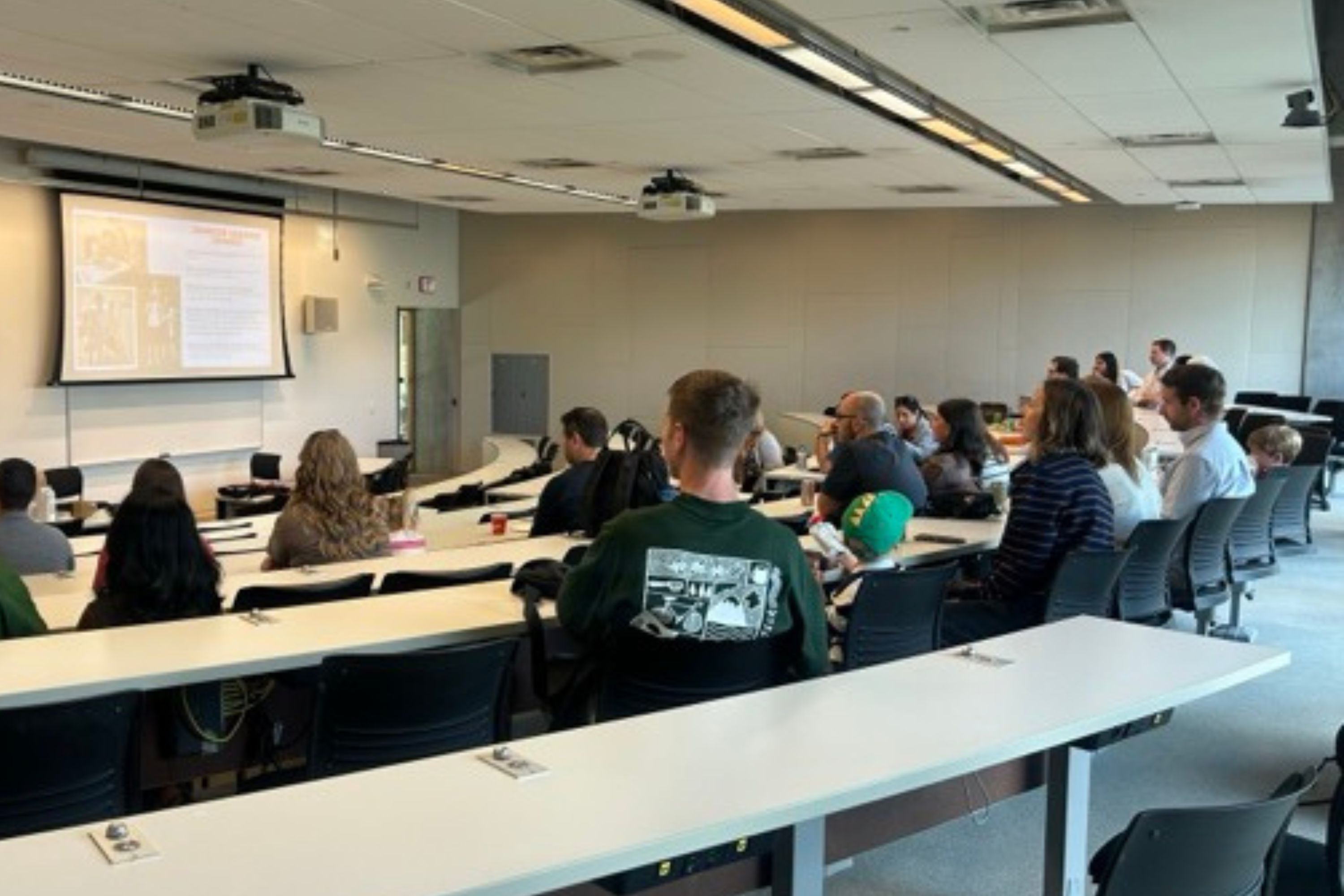 Audience members sit in a tiered classroom facing a projected presentation at the front of the room.