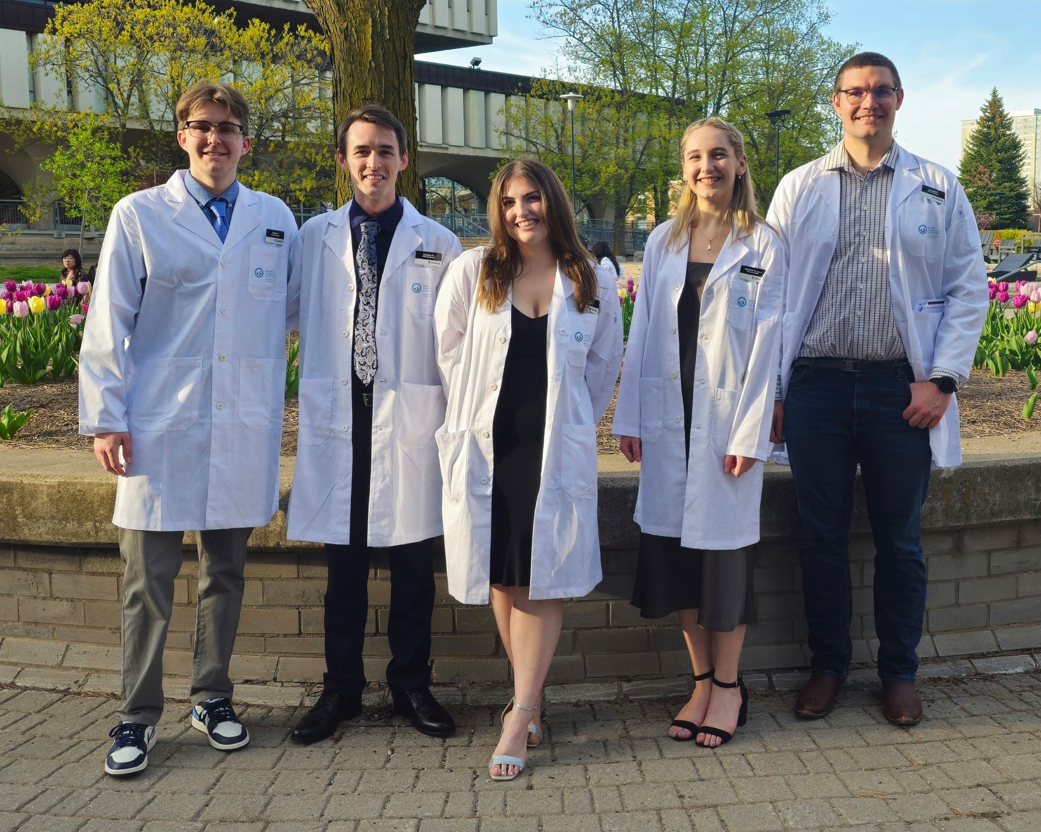 Five people wearing white lab coats stand outdoors in a landscaped campus setting with blooming tulips and leafy trees. They are positioned in a line in front of a low brick wall, with a modern building visible in the background. The group faces the camera, and some wear name tags and formal attire underneath their lab coats.