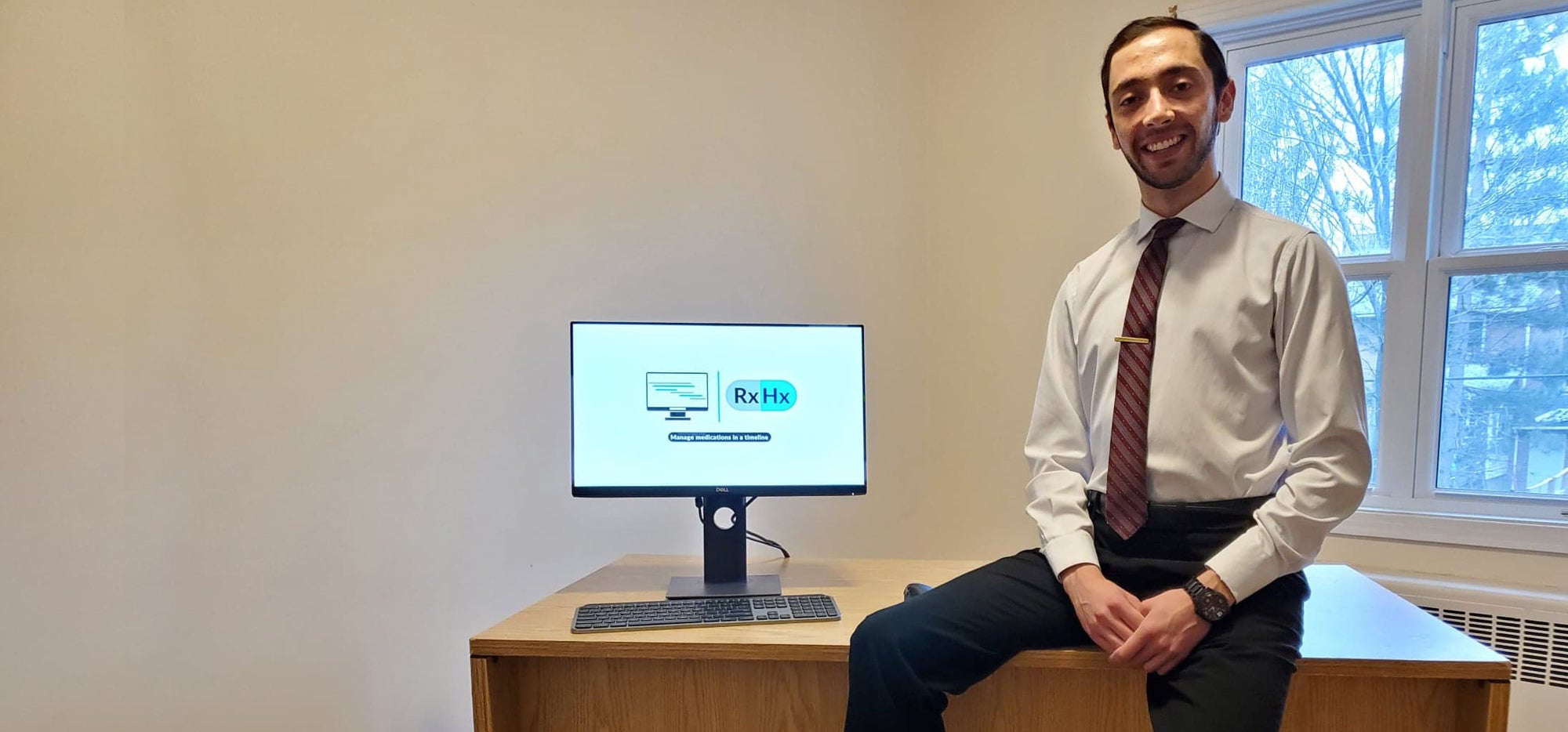 George Daskalakis sitting on desk next to screen showing RxHx logo