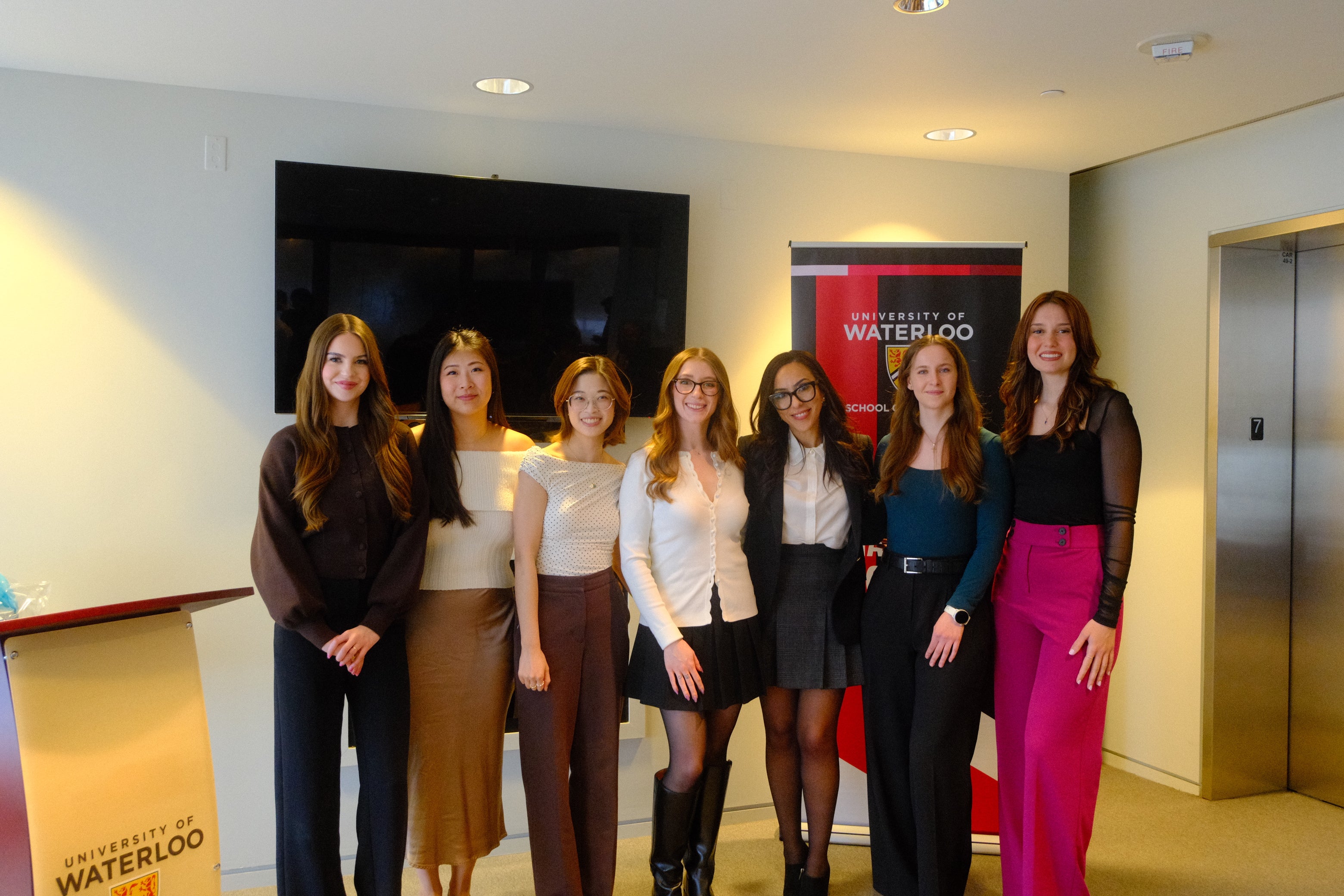 Seven people stand in a row indoors in a well-lit, modern room. A University of Waterloo banner is positioned behind the group, with a wall-mounted television to the left and elevator doors to the right. The people are dressed in professional and semi-formal attire, facing the camera.