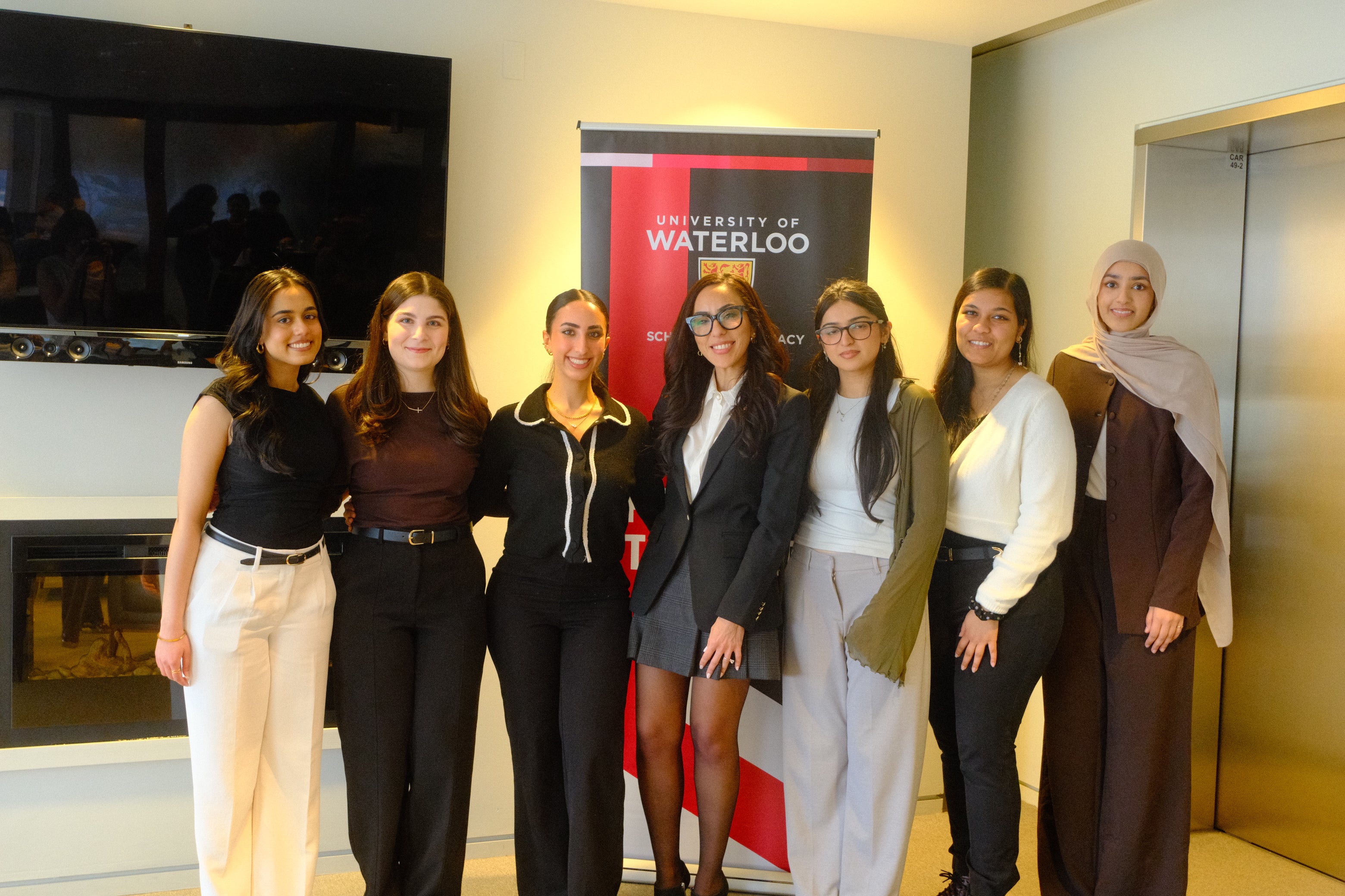 Eight people stand side by side indoors in a bright, modern room. A red-and-black University of Waterloo banner is positioned behind the group, with a wall-mounted television and fireplace visible on the left and closed elevator doors on the right. The group faces the camera, dressed in professional and semi-formal clothing.