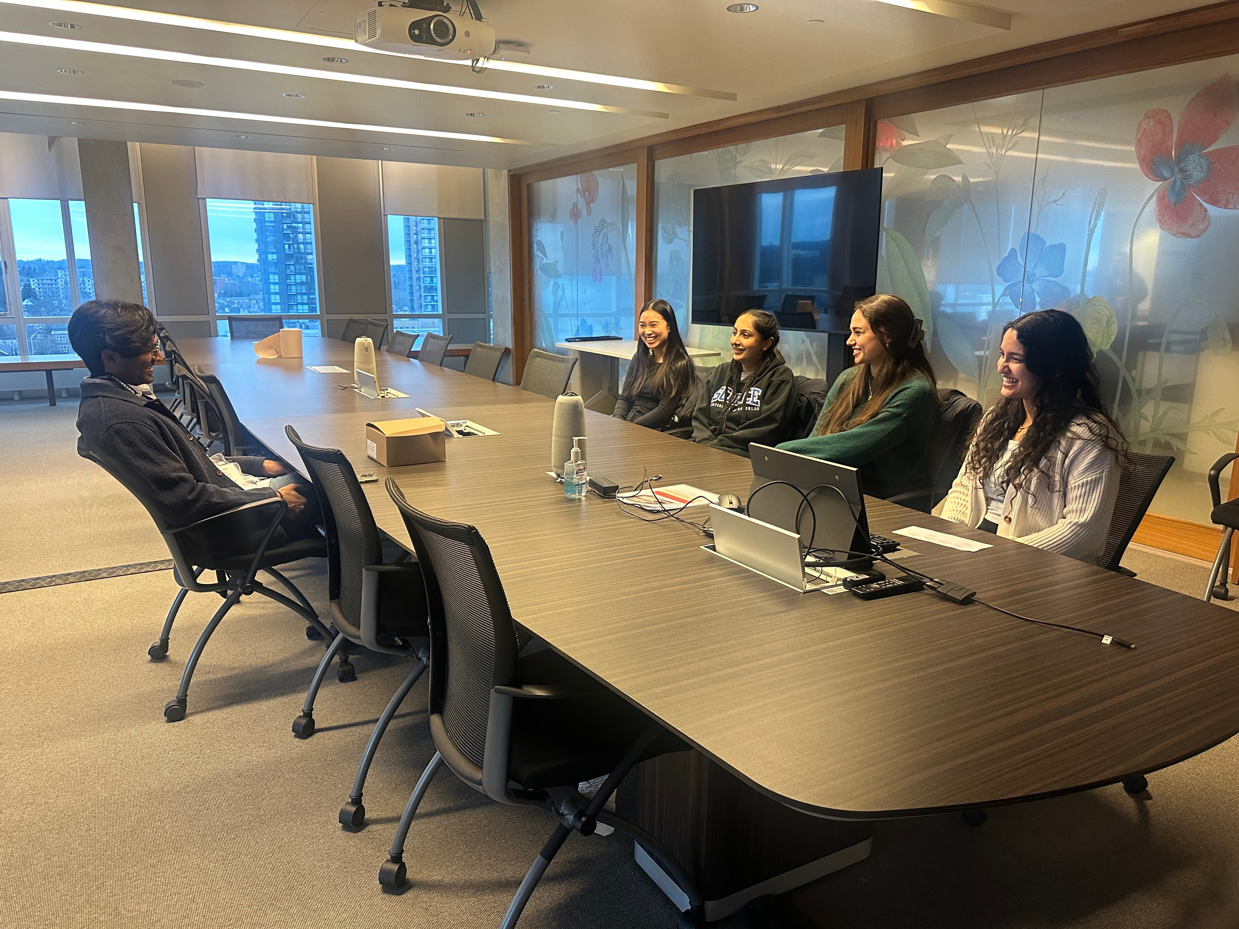 Large conference room with a long rectangular table and rolling office chairs. One adult sits slightly apart at one end of the table, while a small group of adults sit along the opposite side, all oriented toward one another during a discussion. Laptops, cables, a reusable water bottle, and a small box rest on the tabletop. The room features glass walls with floral designs, large windows showing an urban skyline, a ceiling‑mounted projector, and bright overhead lighting, suggesting a formal meeting, intervi
