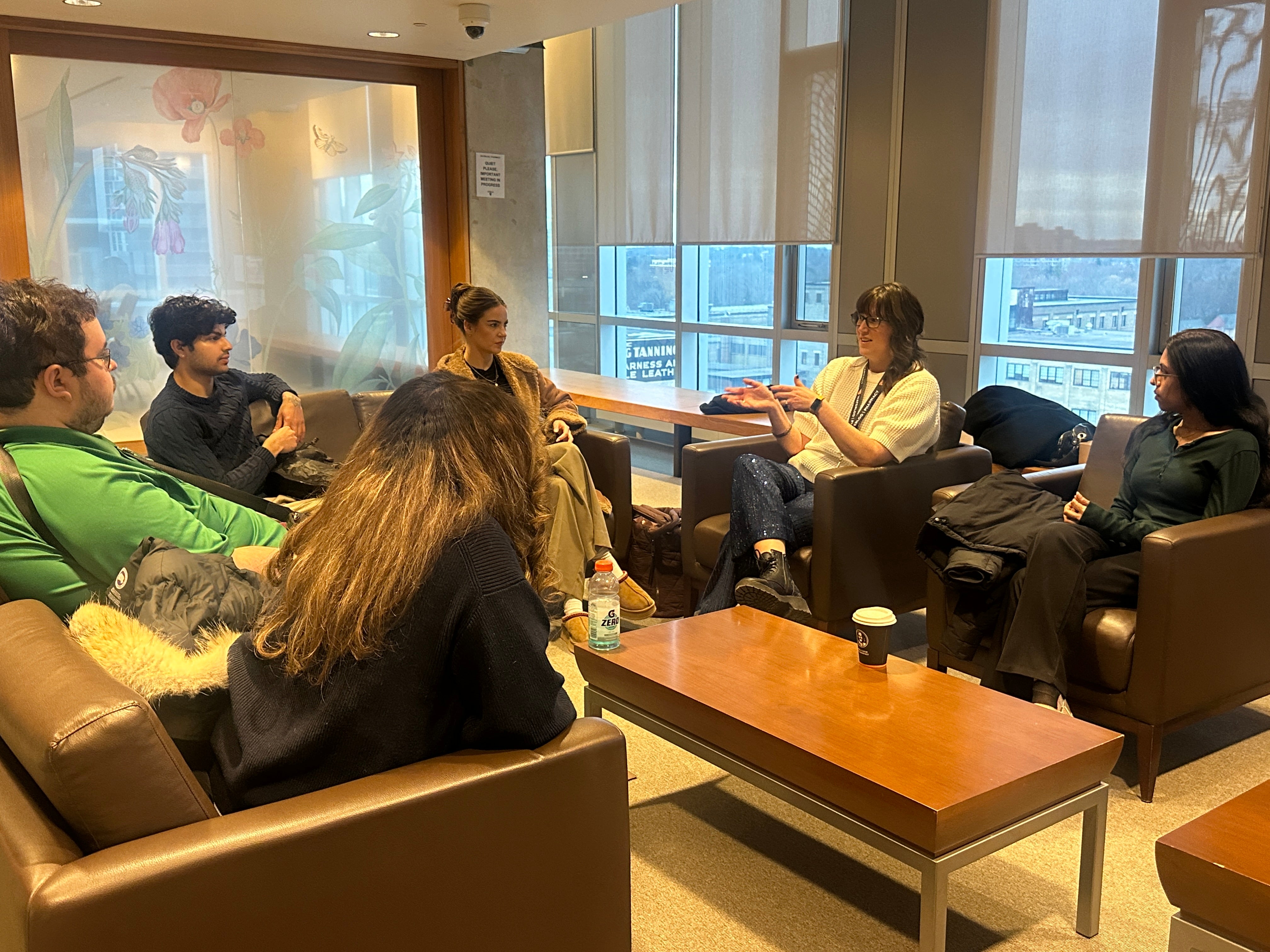 Group of adults seated in armchairs arranged in a circle around a low rectangular coffee table in a lounge-style meeting area. One participant gestures while speaking as others sit facing inward, participating in the discussion. The space has large windows with partially lowered blinds, letting in daylight and revealing nearby buildings outside. Coats, bags, notebooks, a water bottle, and takeaway coffee cups rest on chairs and the table, suggesting an informal group discussion or facilitated conversation i
