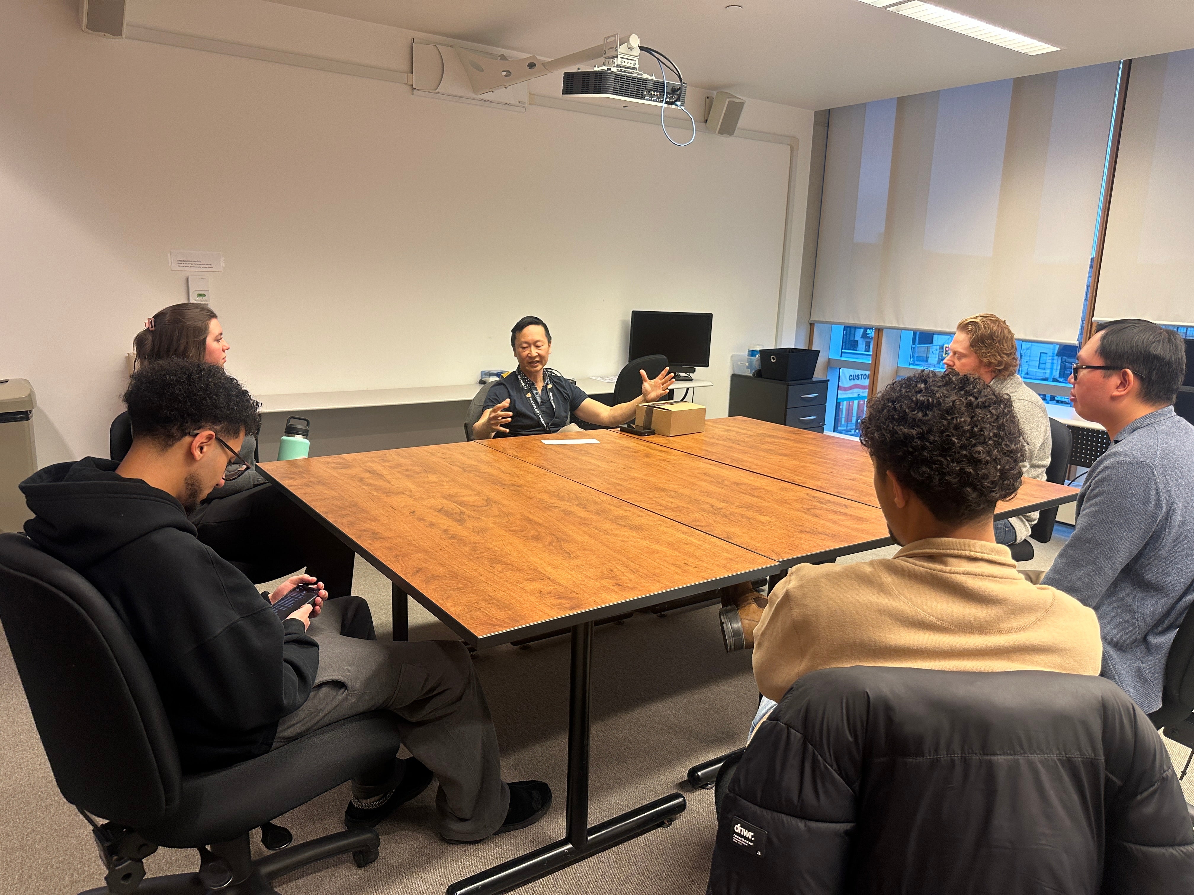 Small group of adults seated around a large rectangular wooden table in a classroom or meeting room. One person seated at the far side of the table speaks while gesturing with both hands, while others sit in rolling office chairs facing inward. The room has light-colored walls, a ceiling-mounted projector, and windows with partially lowered blinds. Personal items such as jackets, notebooks, and a takeaway cup are visible, suggesting a facilitated discussion or small-group meeting.