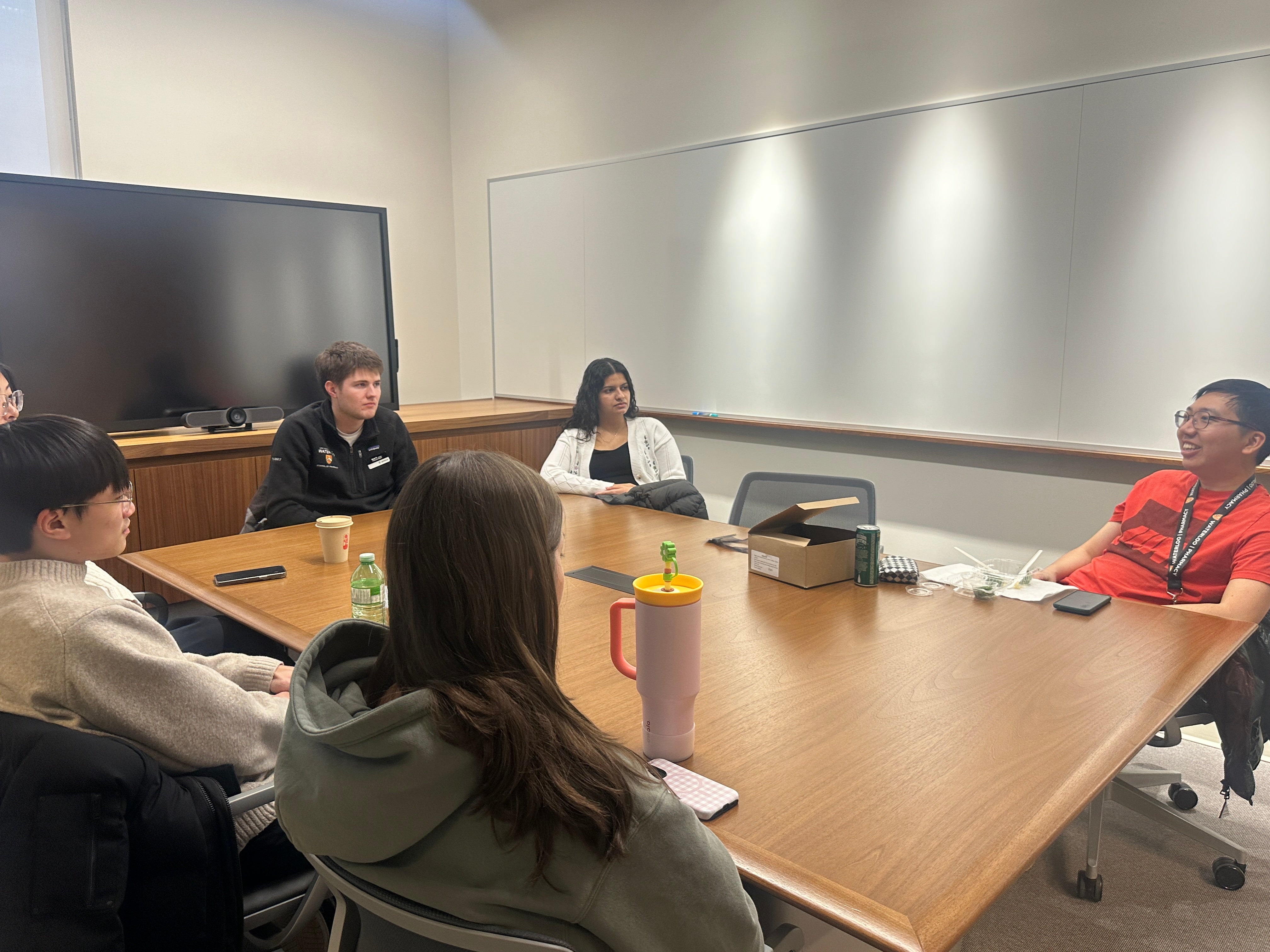 Small group of adults seated around a rectangular wooden conference table in a meeting room. Participants sit in office chairs facing inward, engaged in a discussion, with one person speaking near the far end of the table. Items such as notebooks, reusable water bottles, coffee cups, and a small box are placed on the table. The room features light-colored walls, a large wall‑mounted display screen, and bright overhead lighting, indicating a collaborative meeting or discussion setting.