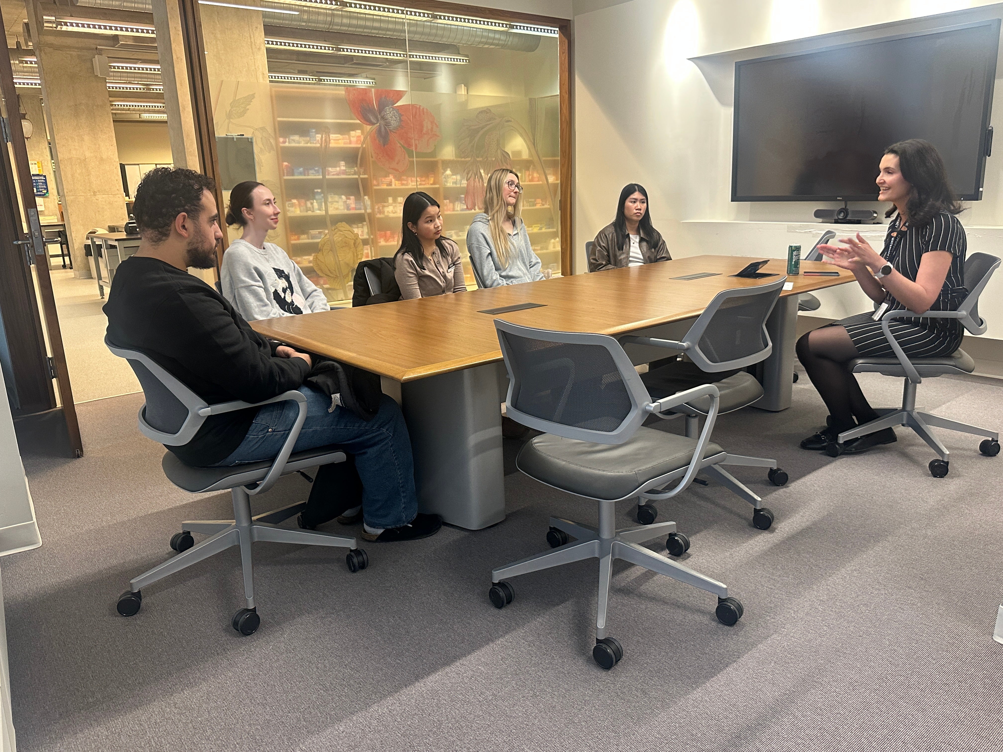 Small group of adults seated around a rectangular wooden meeting table in a conference room. One person speaks while gesturing, and others sit in rolling office chairs facing the speaker. A large wall-mounted display screen, glass walls, and a visible hallway or open workspace appear in the background. Personal items, including notebooks and a drink can, sit on the table, indicating a collaborative meeting or discussion setting.