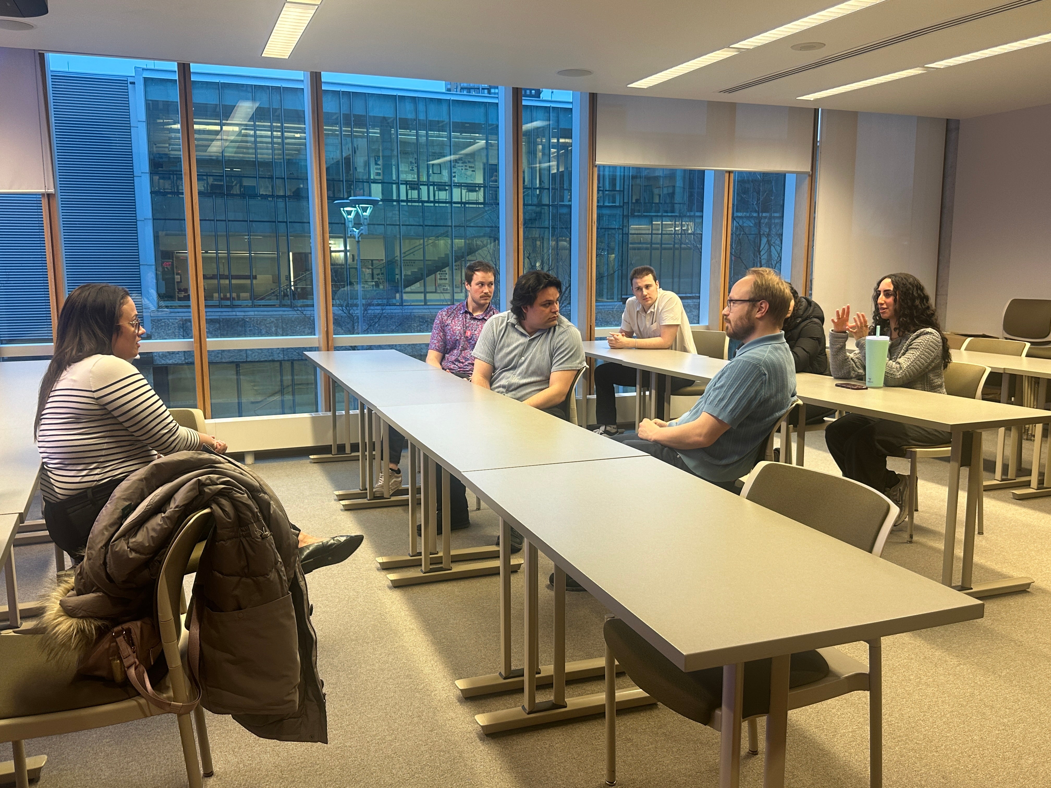 Medium-sized classroom with rows of rectangular tables facing a seated speaker at the front of the room. Several adults sit at the tables, oriented toward the speaker, who is seated on a stool near a podium with a computer monitor. The room features neutral-colored walls, overhead lighting, an exposed ceiling with ducts, a wall clock, and windows along one side. Personal items, including a reusable drink can on the floor, are visible, suggesting a lecture, talk, or facilitated discussion.