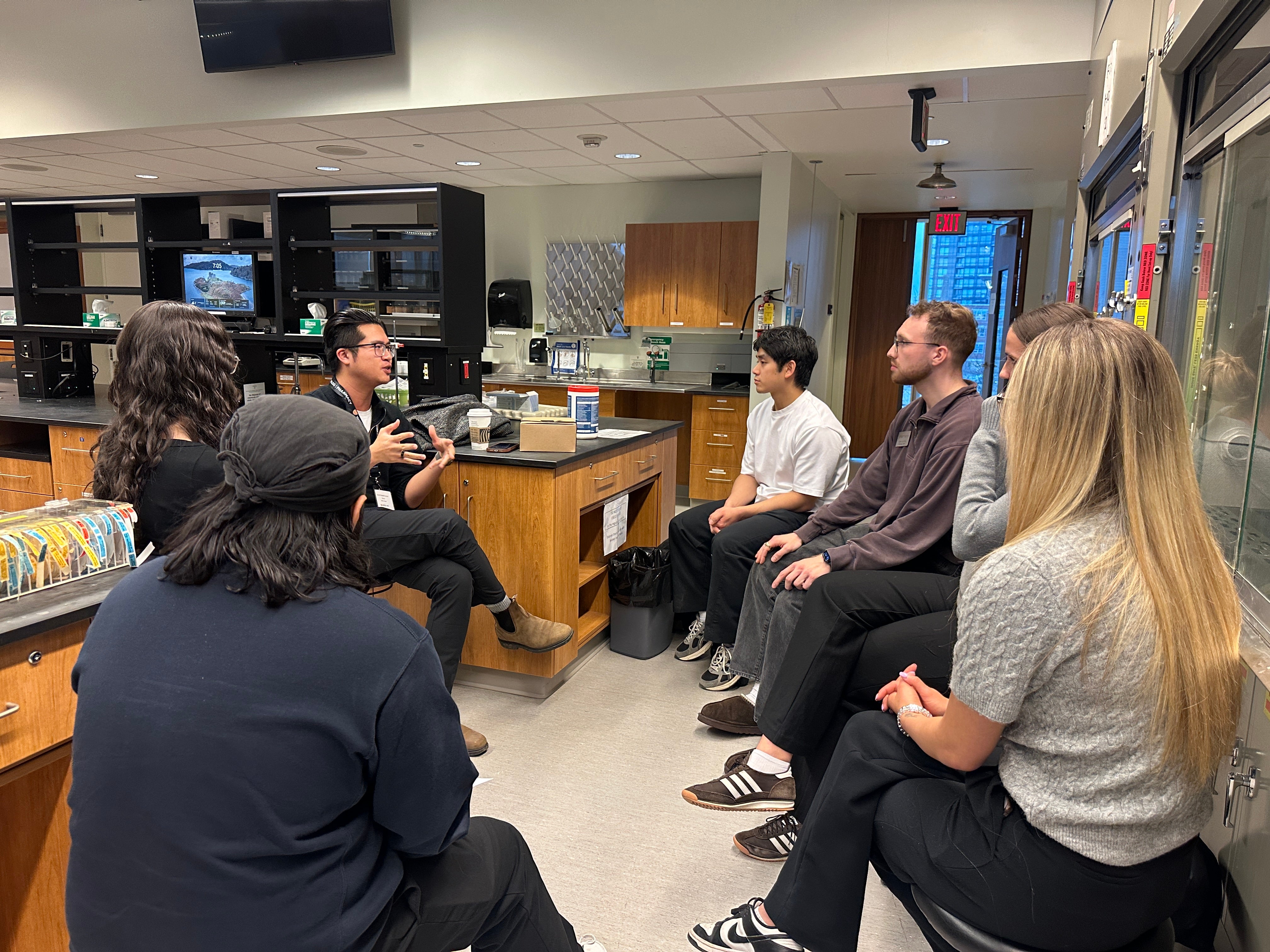 Small group of adults seated in a semicircle inside a laboratory or teaching lab, participating in a discussion. One person gestures while speaking near a central lab bench with cabinets, equipment, and supplies, while others sit on stools along the walls, wearing casual clothing and lanyards under bright indoor lighting.