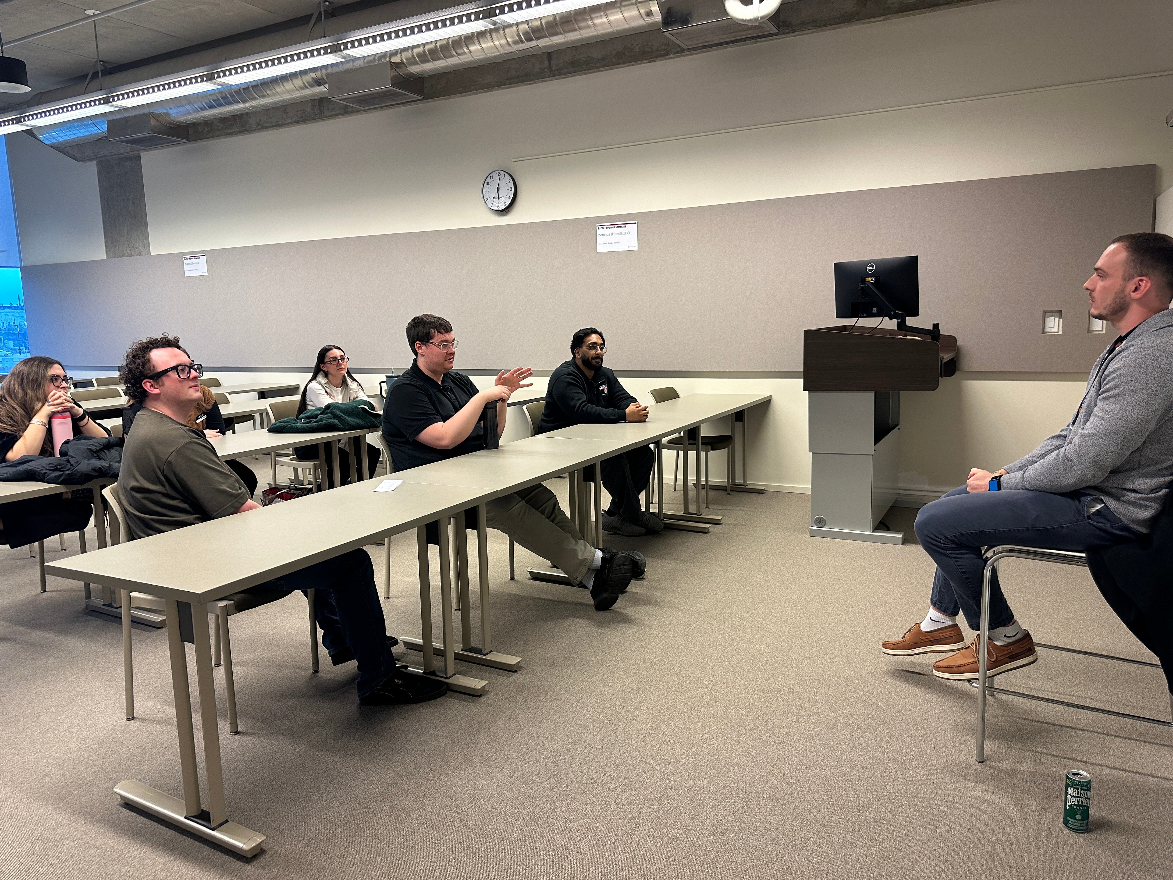 Medium-sized classroom with rows of rectangular tables facing a seated speaker at the front of the room. Several adults sit at the tables, oriented toward the speaker, who is seated on a stool near a podium with a computer monitor. The room features neutral-colored walls, overhead lighting, an exposed ceiling with ducts, a wall clock, and windows along one side. Personal items, including a reusable drink can on the floor, are visible, suggesting a lecture, talk, or facilitated discussion.