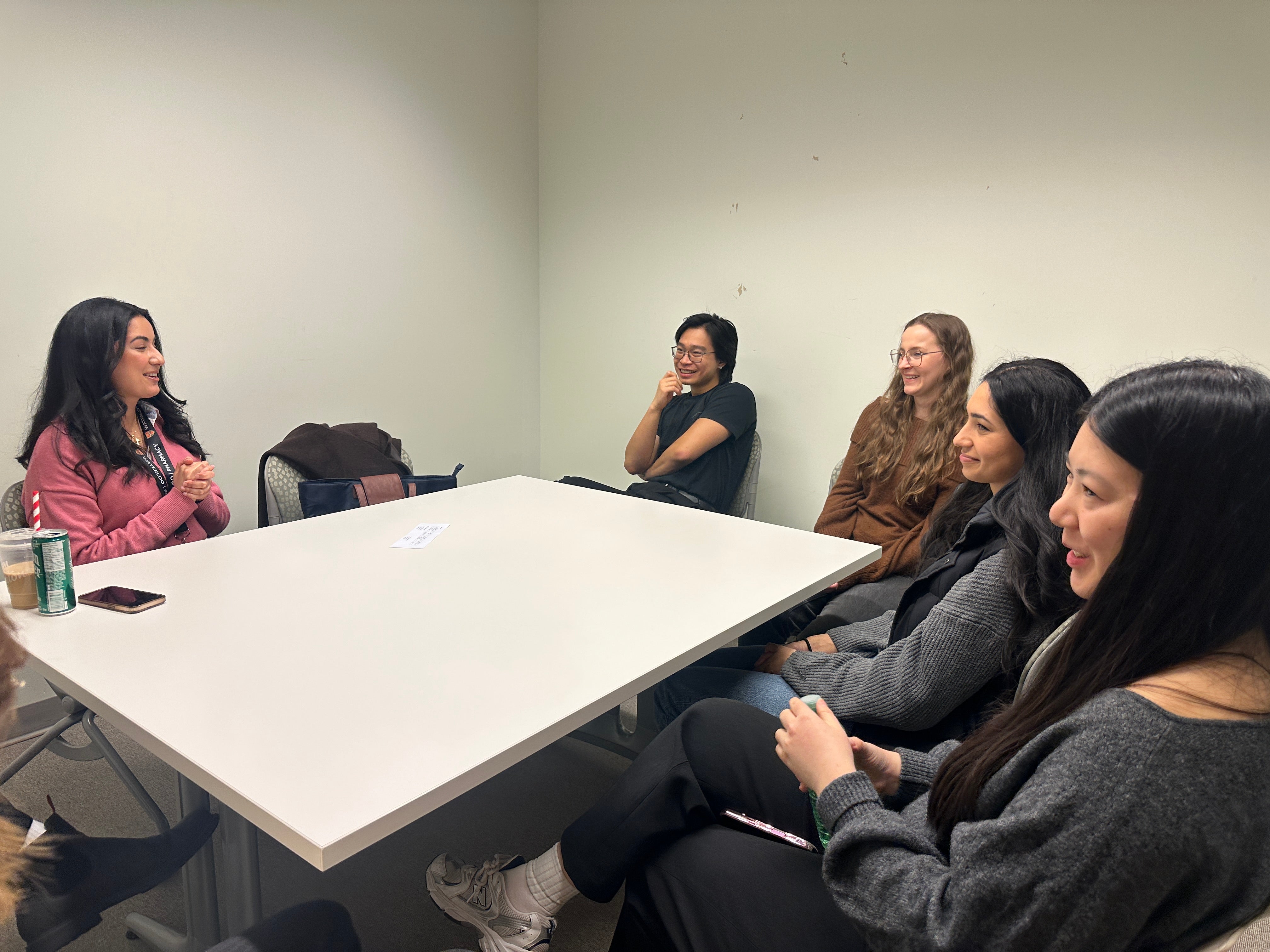 Small group of adults seated around a rectangular white table in a plain meeting room, engaged in conversation. One person speaks with hands clasped while others sit along the table edges, facing toward the speaker. Personal items such as notebooks, phones, a reusable cup, and outerwear are visible on and around the table. The room has light-colored walls, simple chairs, and even overhead lighting, suggesting an informal discussion or small-group meeting.