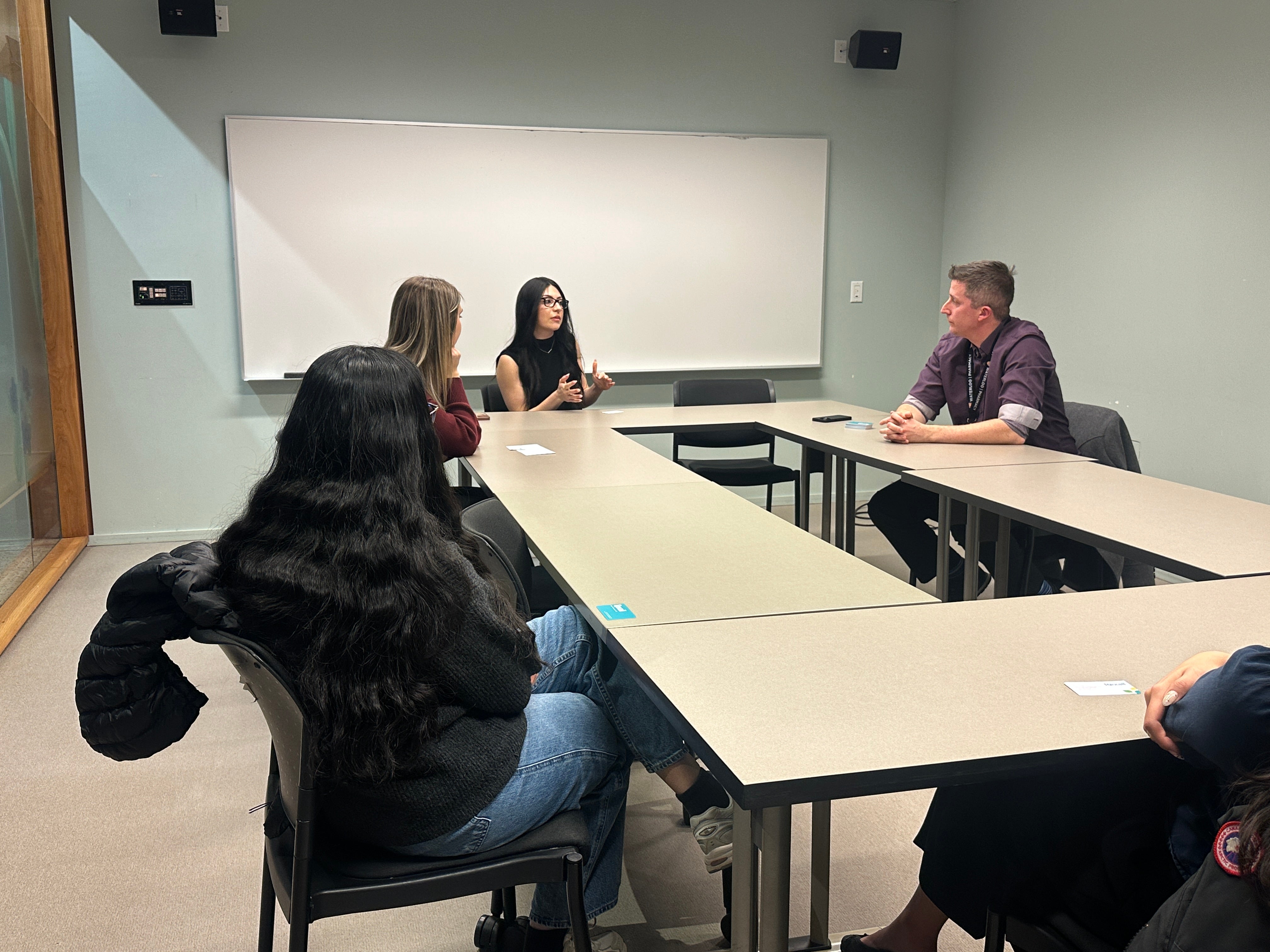 Small group of adults seated around rectangular tables arranged in a U-shape in a classroom or meeting room. One person at the far side of the tables appears to be speaking and gesturing, while others sit and listen. The room has light-colored walls, a whiteboard mounted at the front, and simple chairs. Personal items such as notebooks and jackets are visible, and the space is evenly lit, suggesting a discussion-based meeting or workshop.
