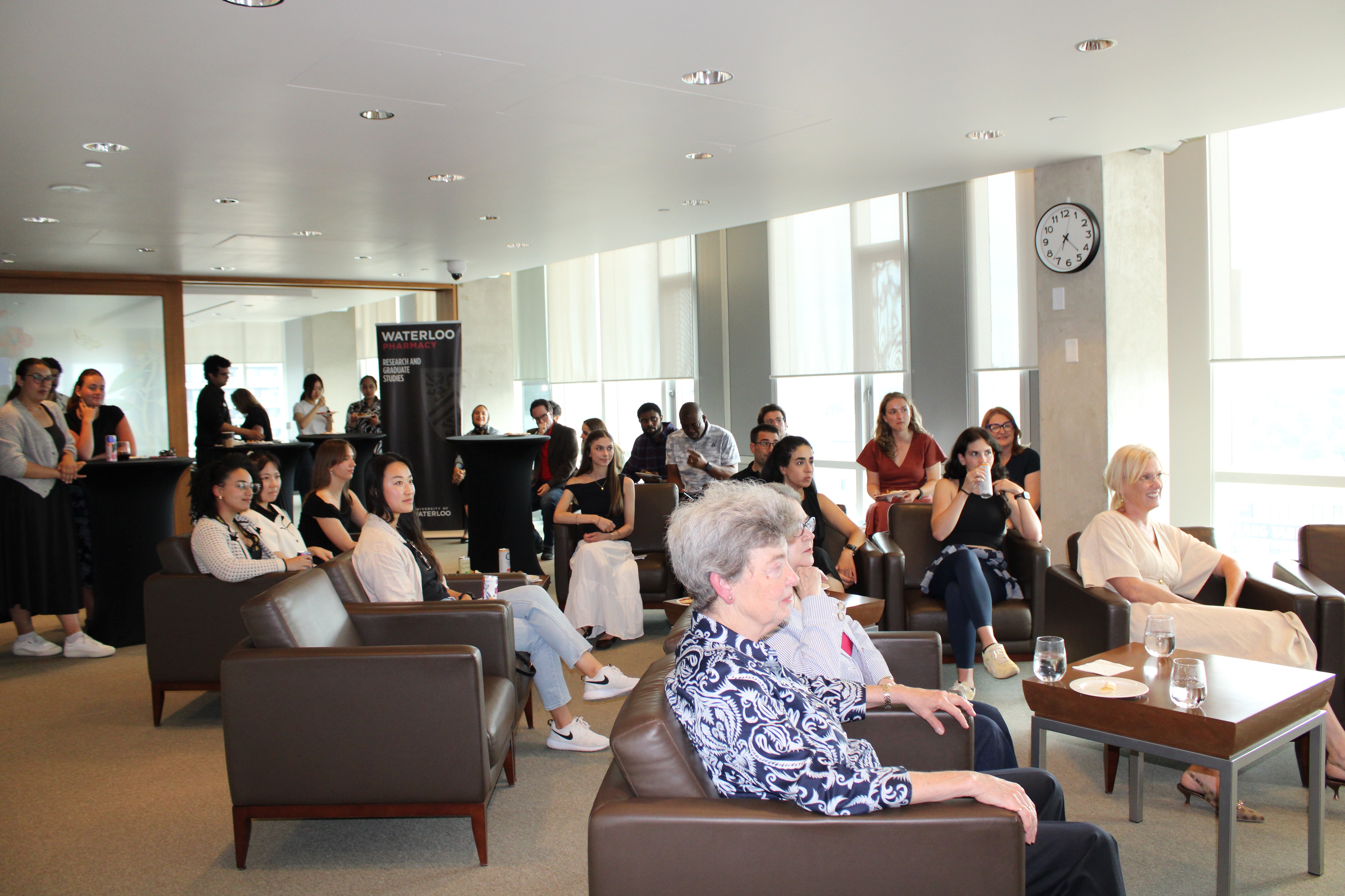 Audience seated in a bright campus lounge, watching a presentation near a Waterloo Pharmacy banner.