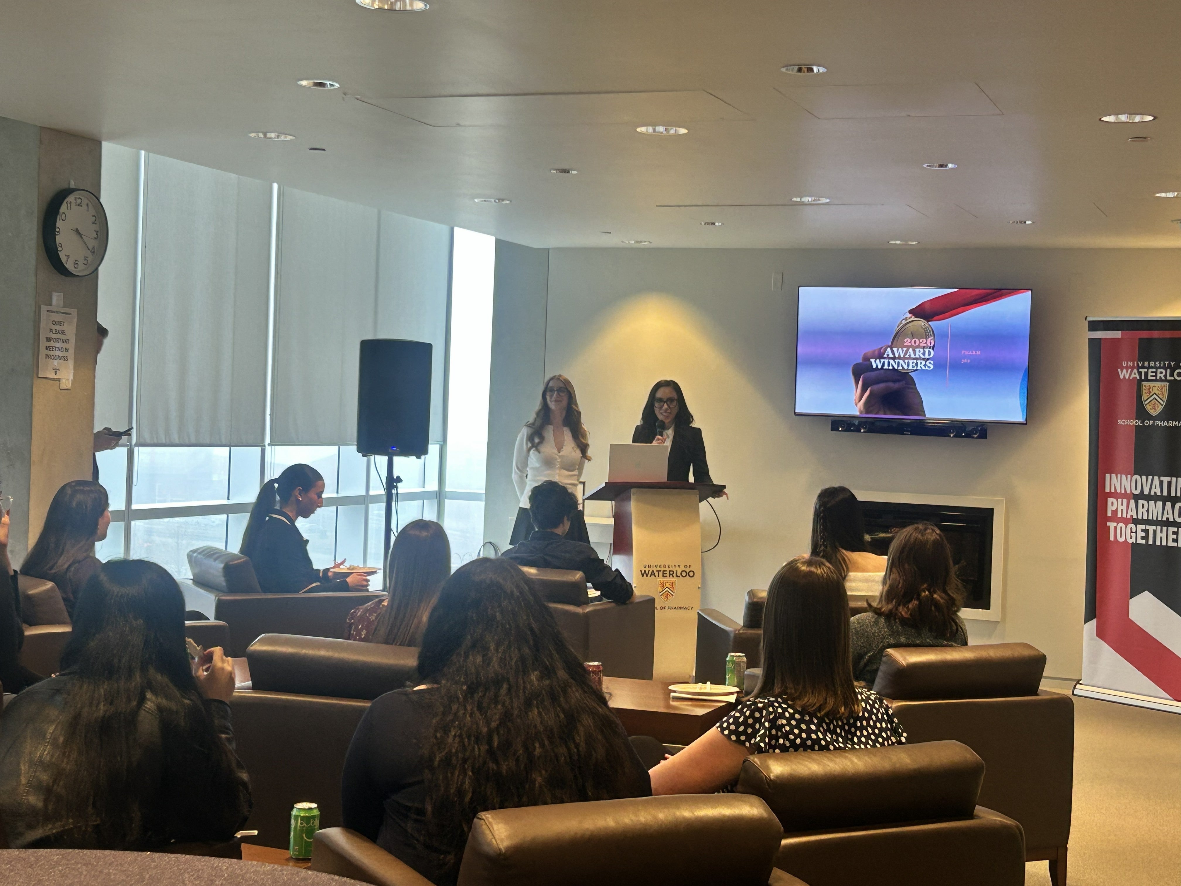 Audience seated in a modern lounge-style room listens as two people stand behind a podium with a University of Waterloo logo, presenting in front of a screen that reads “Award Winners 2023,” with banners and large windows visible in the background.