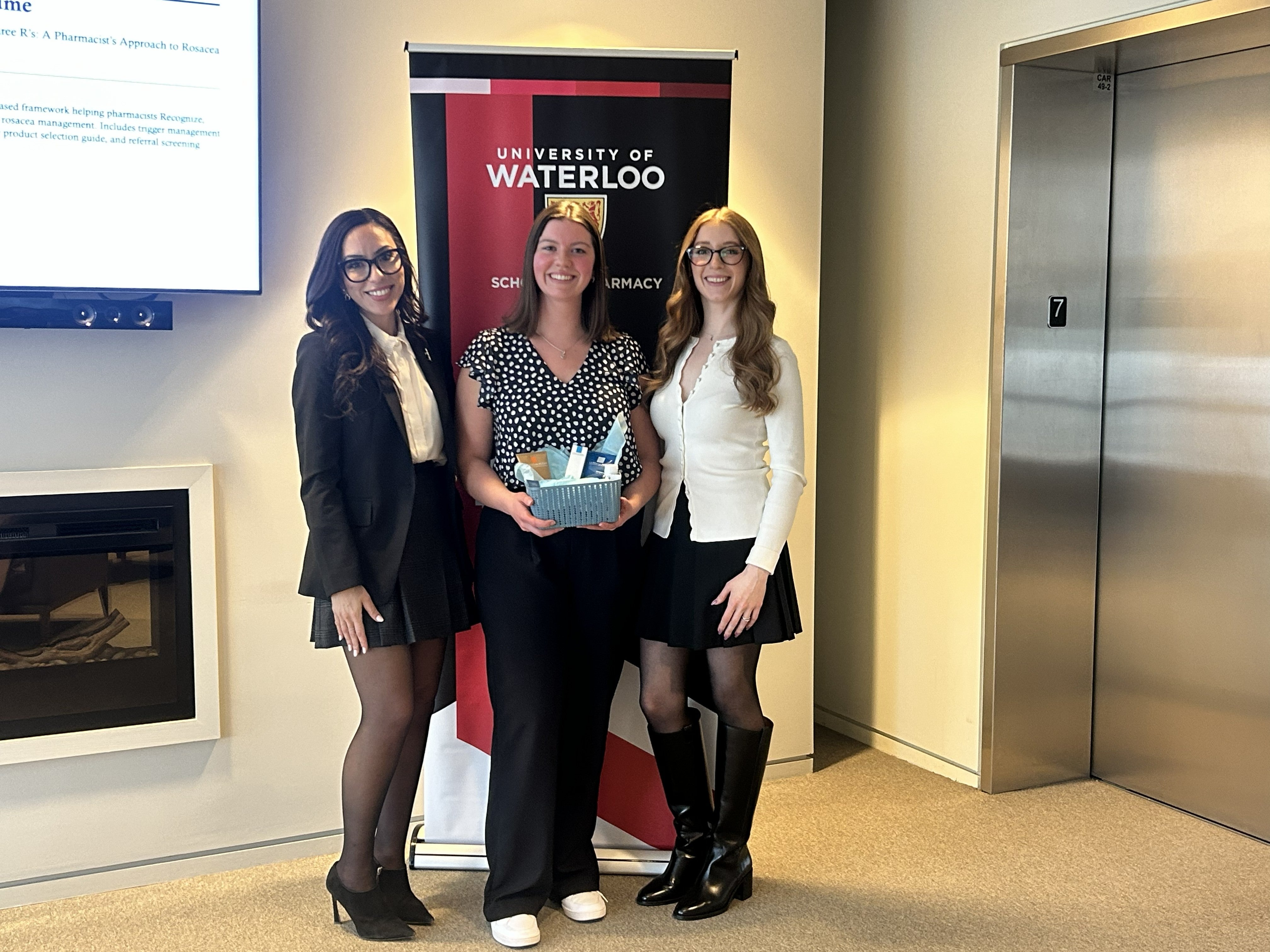 Three people stand side by side posing with a small gift basket in front of a University of Waterloo School of Pharmacy banner inside a bright indoor space, with an elevator and wall-mounted screen visible in the background.