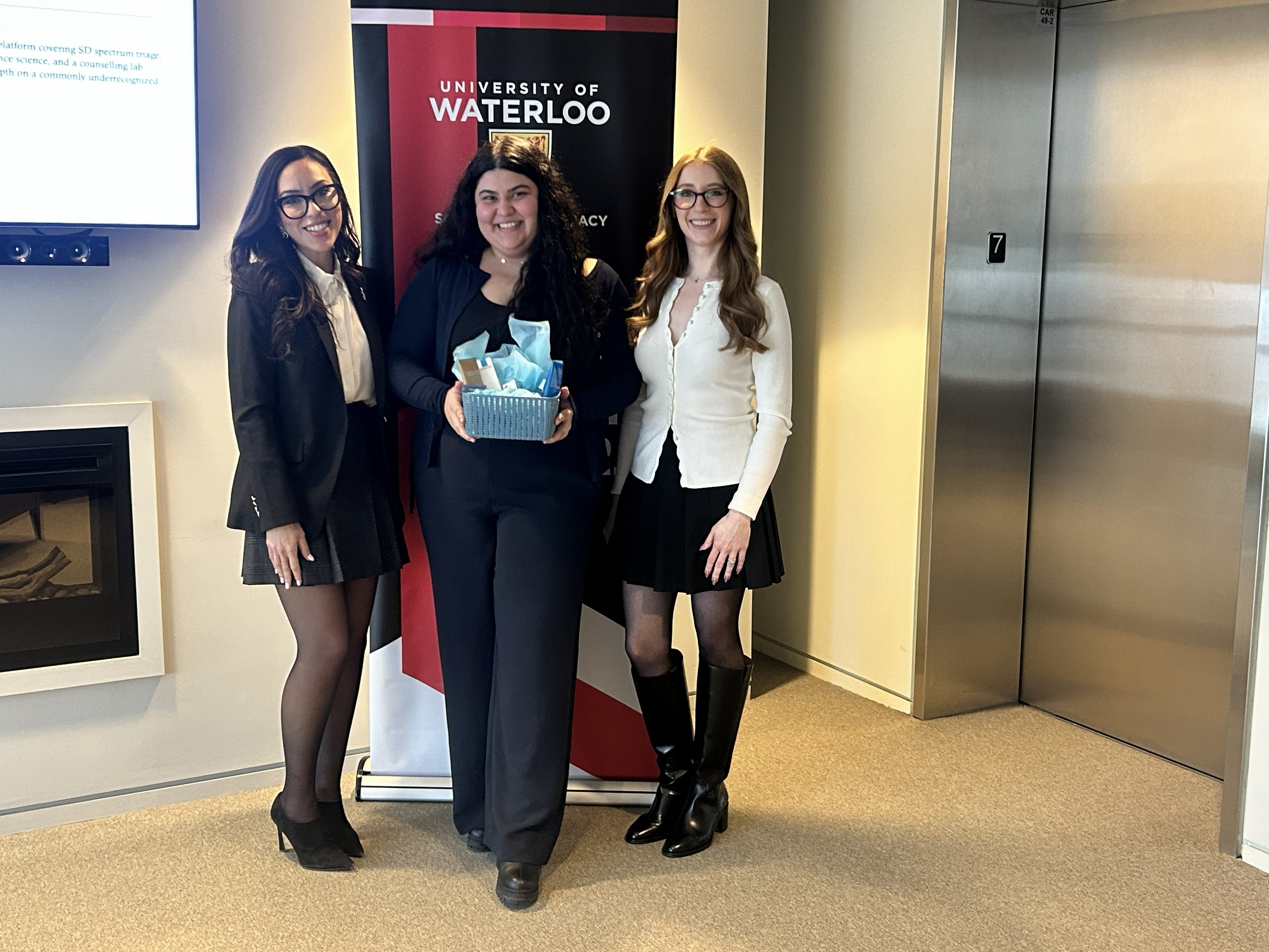 Three people stand side by side posing with a small gift basket in front of a University of Waterloo School of Pharmacy banner inside a bright indoor space, with an elevator and wall-mounted screen visible in the background.
