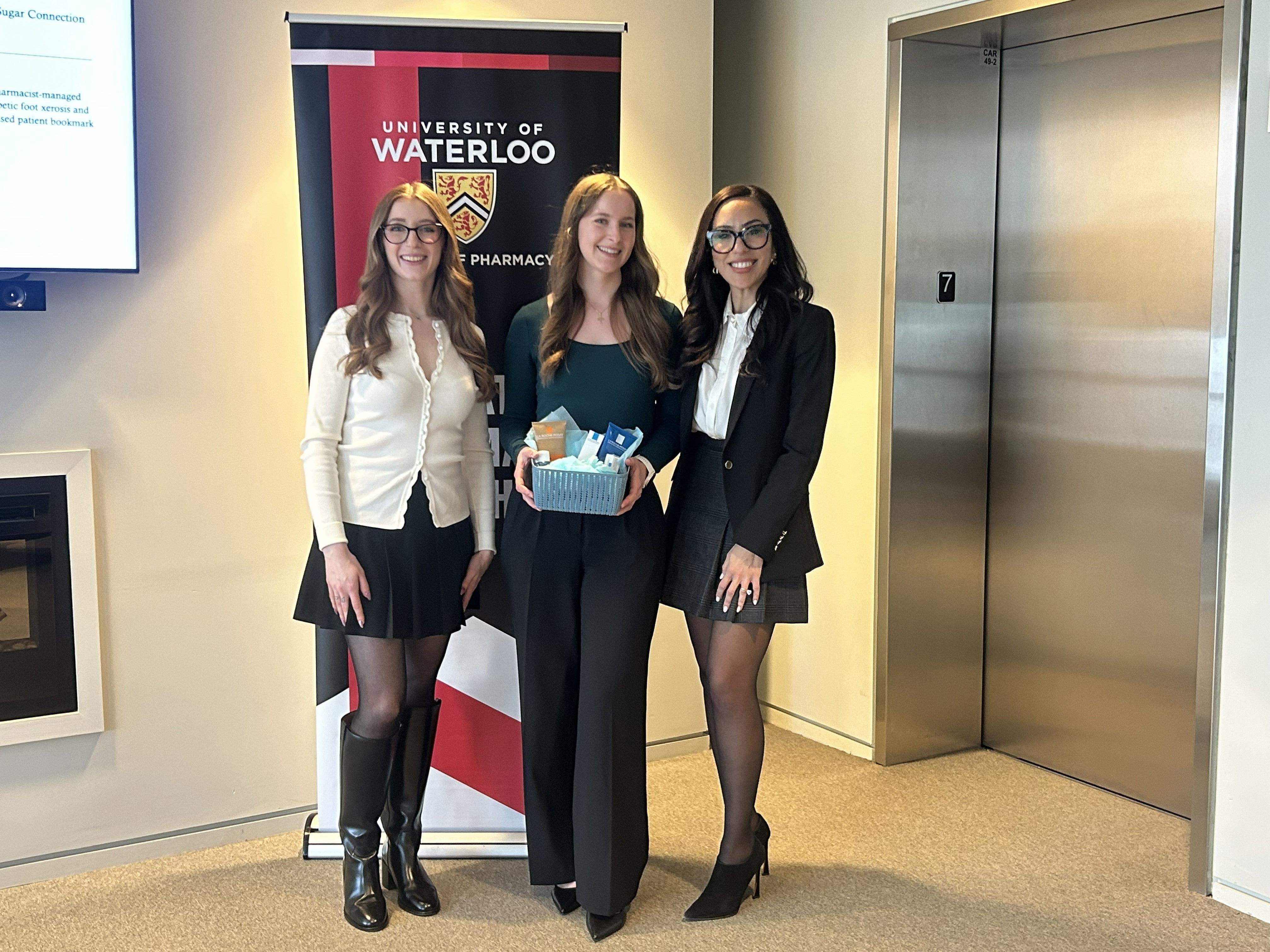 Three people stand side by side posing with a small gift basket in front of a University of Waterloo School of Pharmacy banner inside a bright indoor space, with an elevator and wall-mounted screen visible in the background.