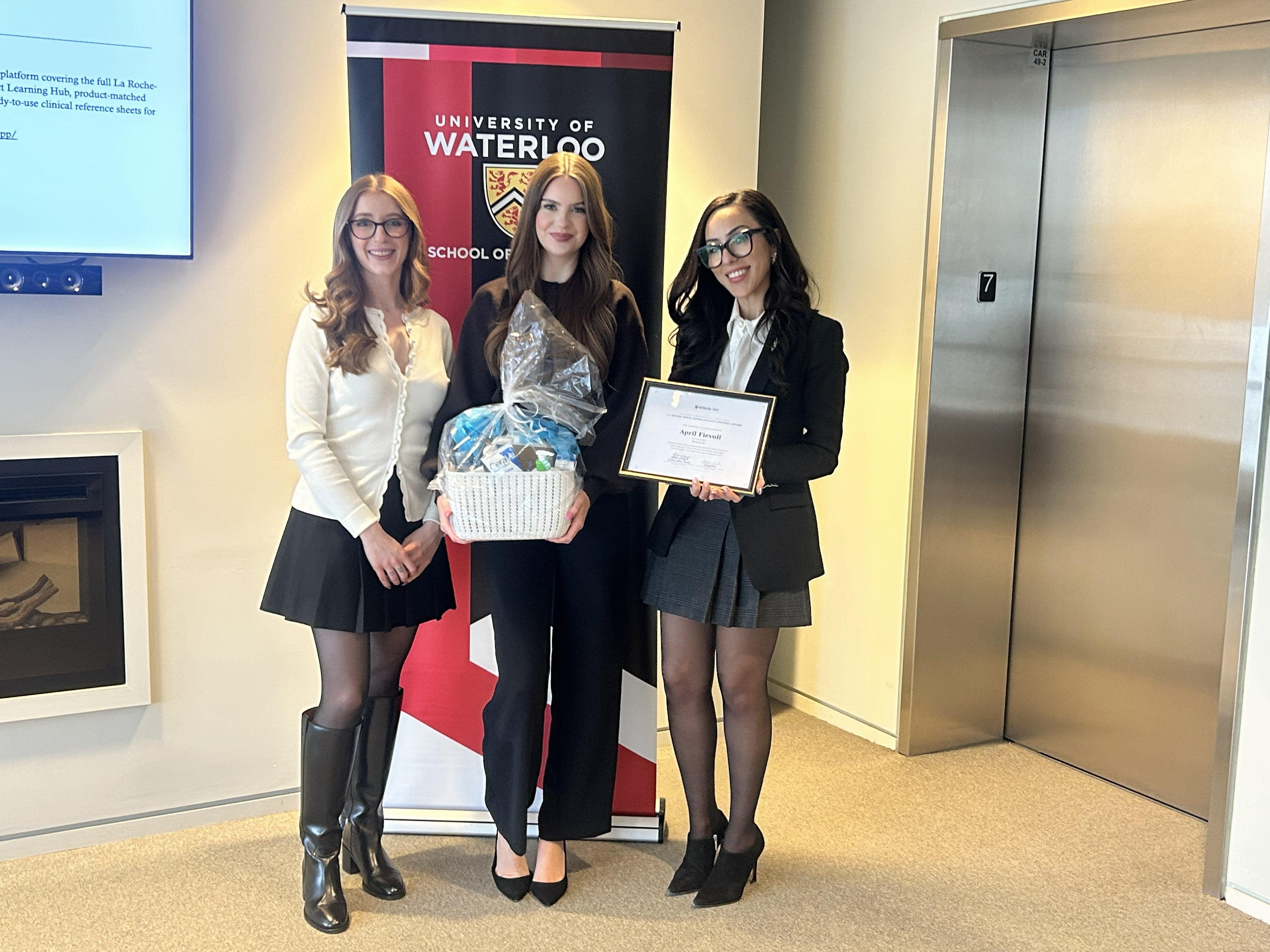 Three people stand side by side posing with a small gift basket in front of a University of Waterloo School of Pharmacy banner inside a bright indoor space, with an elevator and wall-mounted screen visible in the background.