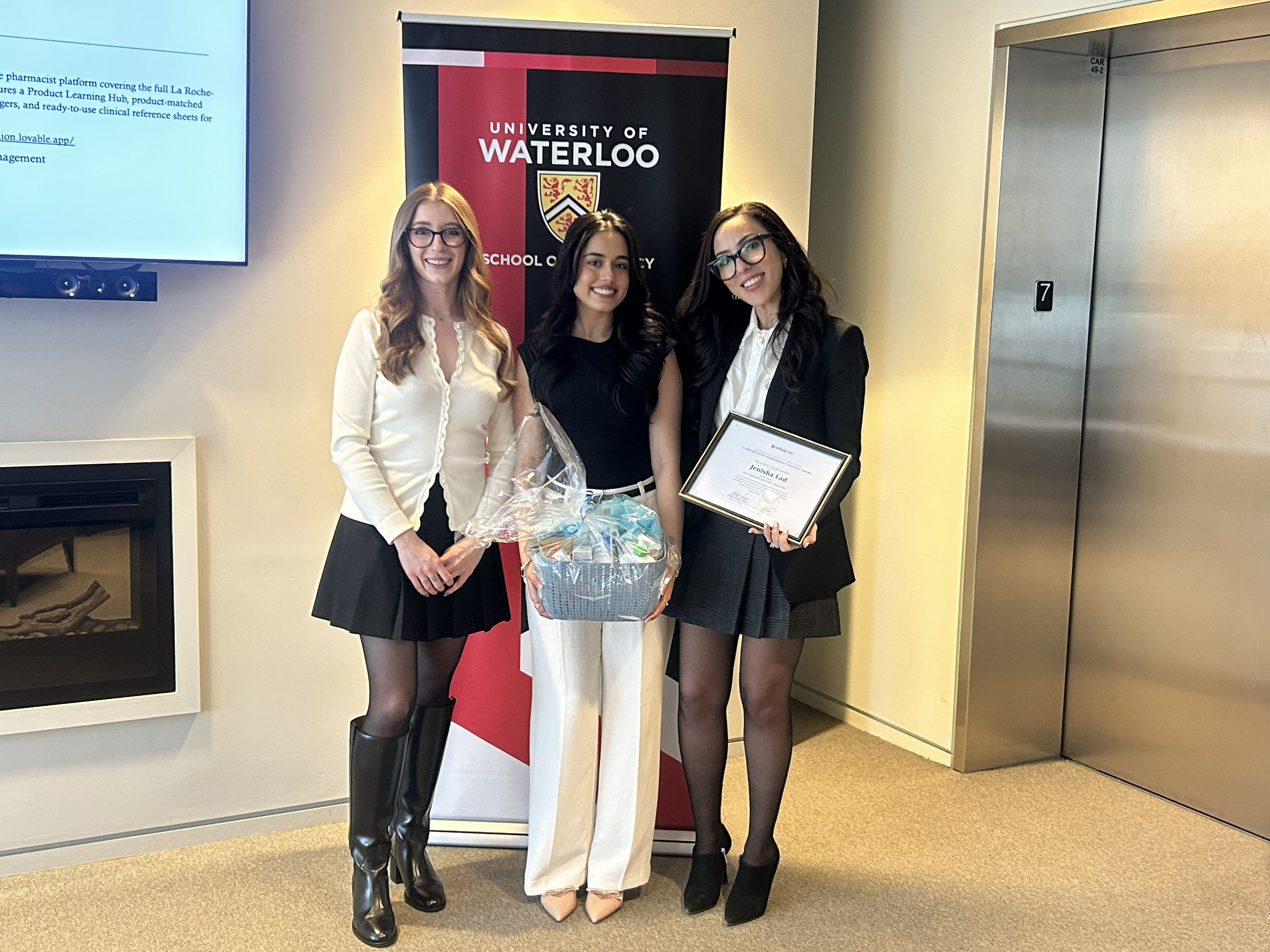 Three people stand side by side posing with a small gift basket in front of a University of Waterloo School of Pharmacy banner inside a bright indoor space, with an elevator and wall-mounted screen visible in the background.