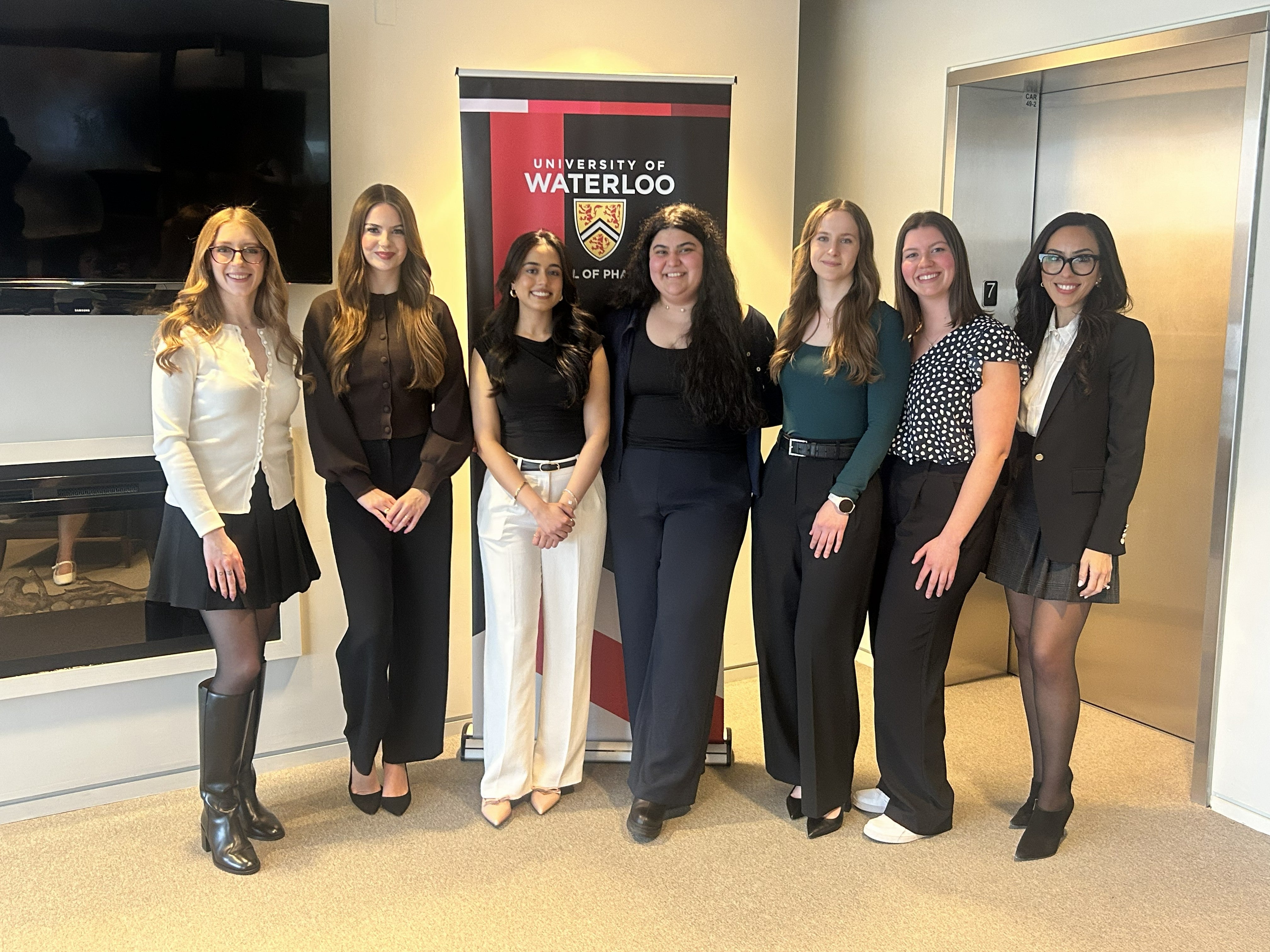 A group of eight people stand in a row posing for a photo indoors in front of a University of Waterloo School of Pharmacy banner, dressed in business or business-casual attire, with a wall-mounted screen on one side and an elevator on the other.