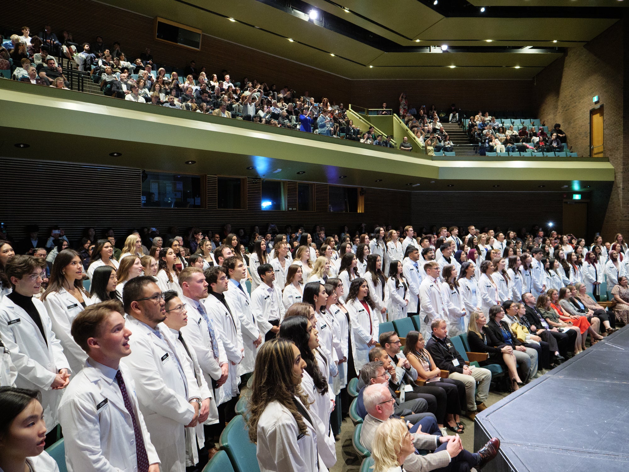 Group of pharmacy students in white coats standing in an auditorium during a ceremony, with audience members seated in front.