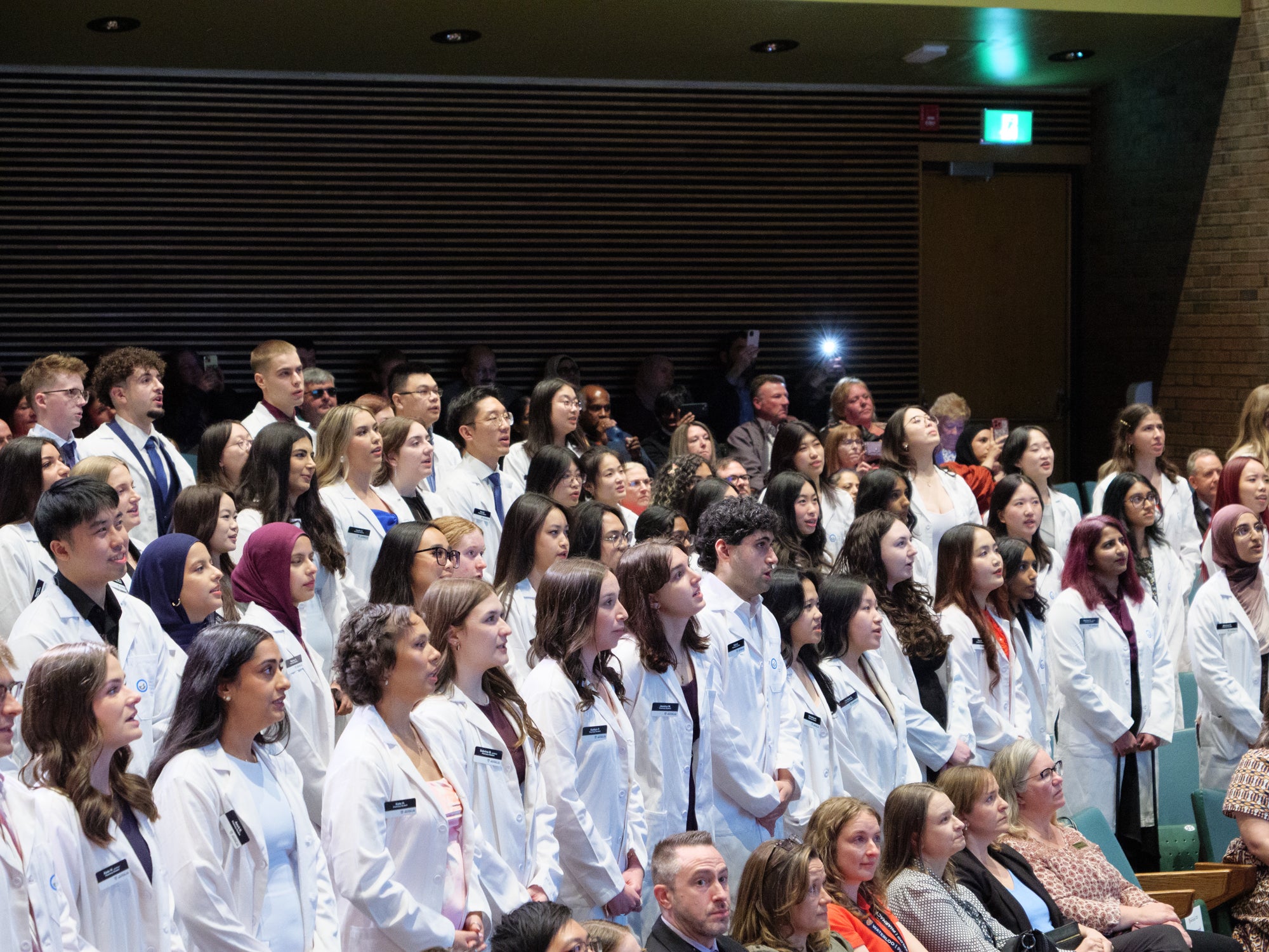 Pharmacy students in white coats standing together during a ceremony in an auditorium.