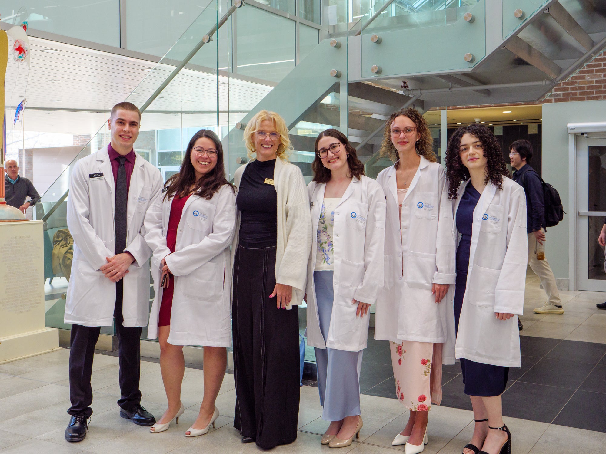 Small group of pharmacy students in white coats posing with a faculty member in a bright campus atrium.