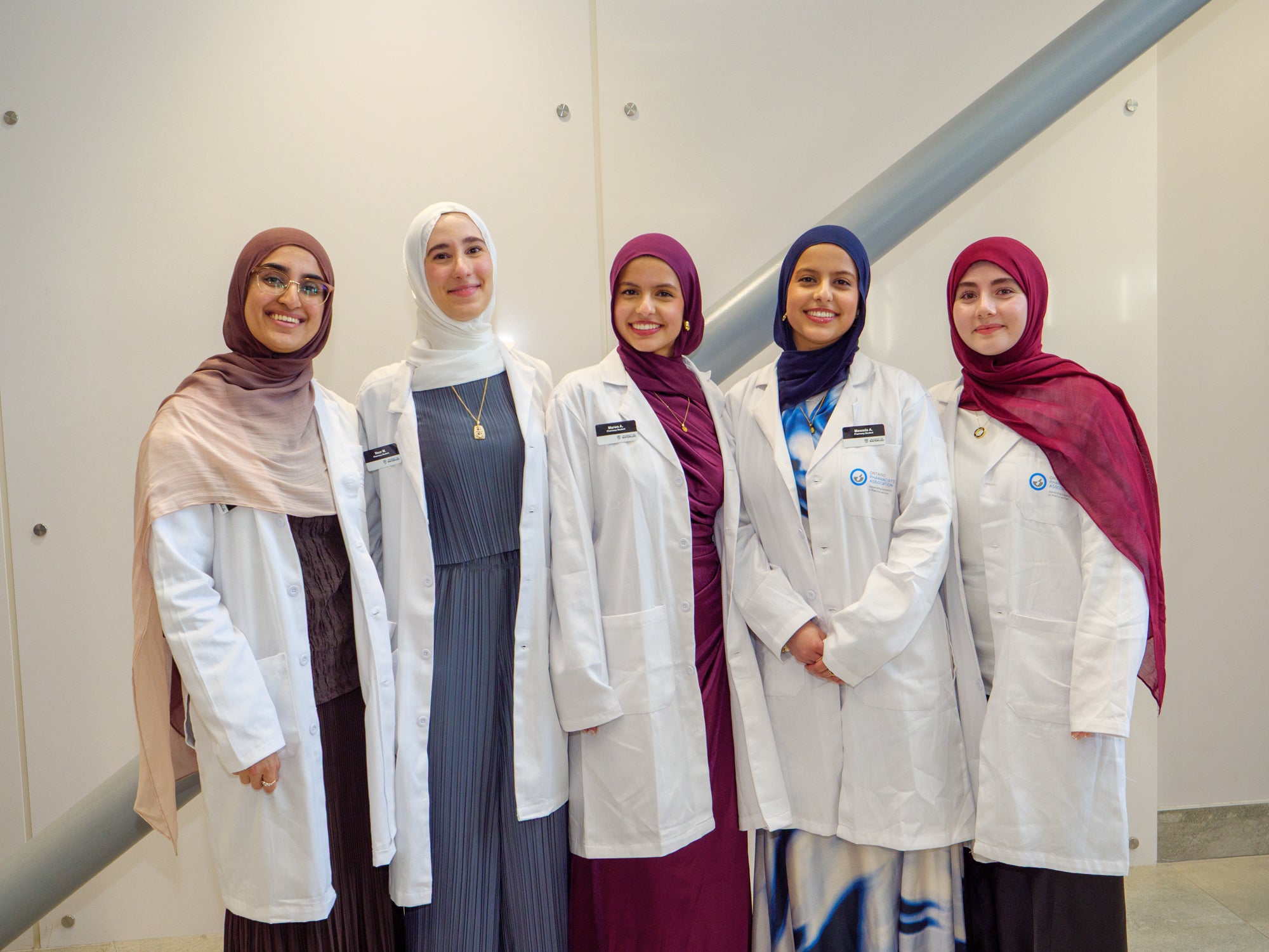 Five pharmacy students in white coats and headscarves posing together indoors.