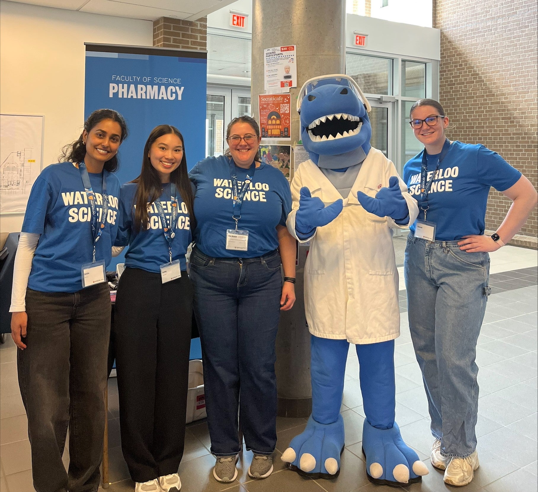 Five people wearing blue “Waterloo Science” shirts stand smiling beside a blue shark mascot in a white lab coat at a Faculty of Science Pharmacy booth.
