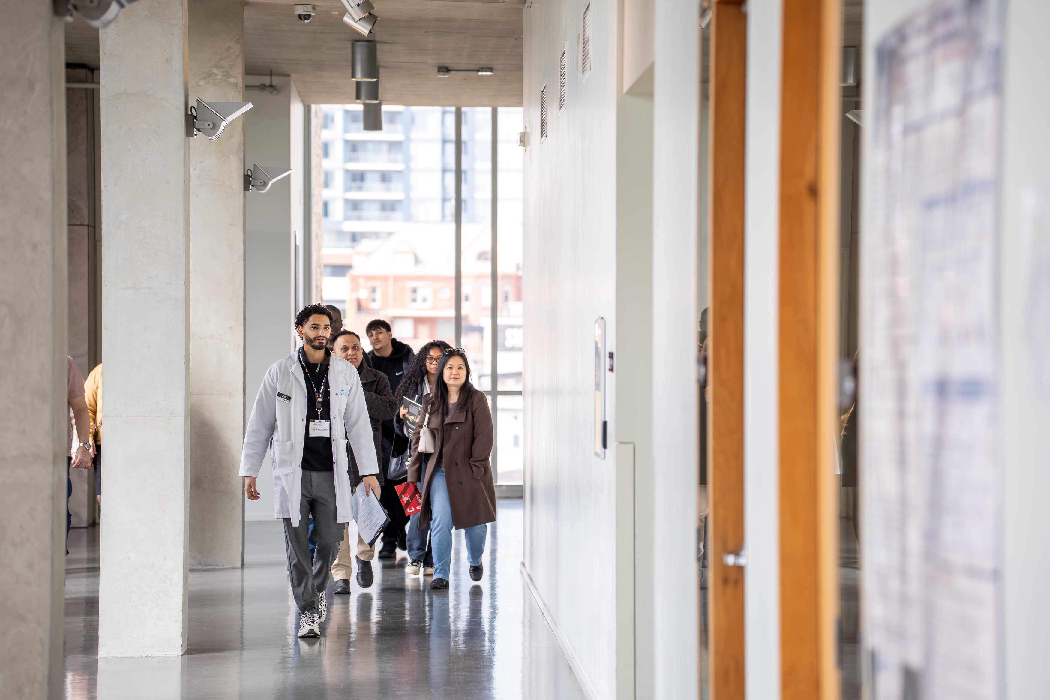 A group of people walk down a bright campus hallway, led by a person wearing a white lab coat.
