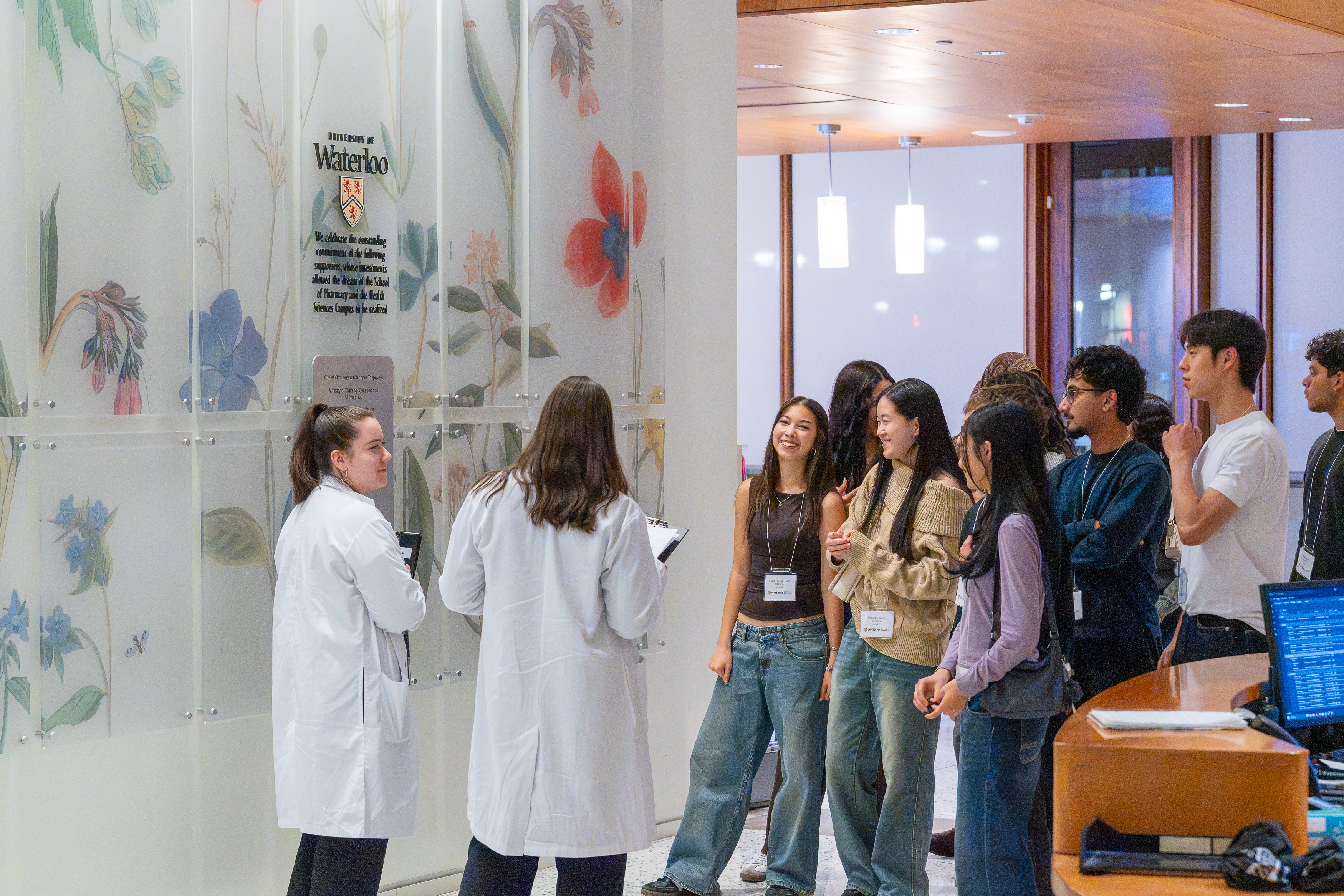 A group of attendees wearing name badges gather near two people in white lab coats in a University of Waterloo building, with a floral wall display and University of Waterloo crest in the background.