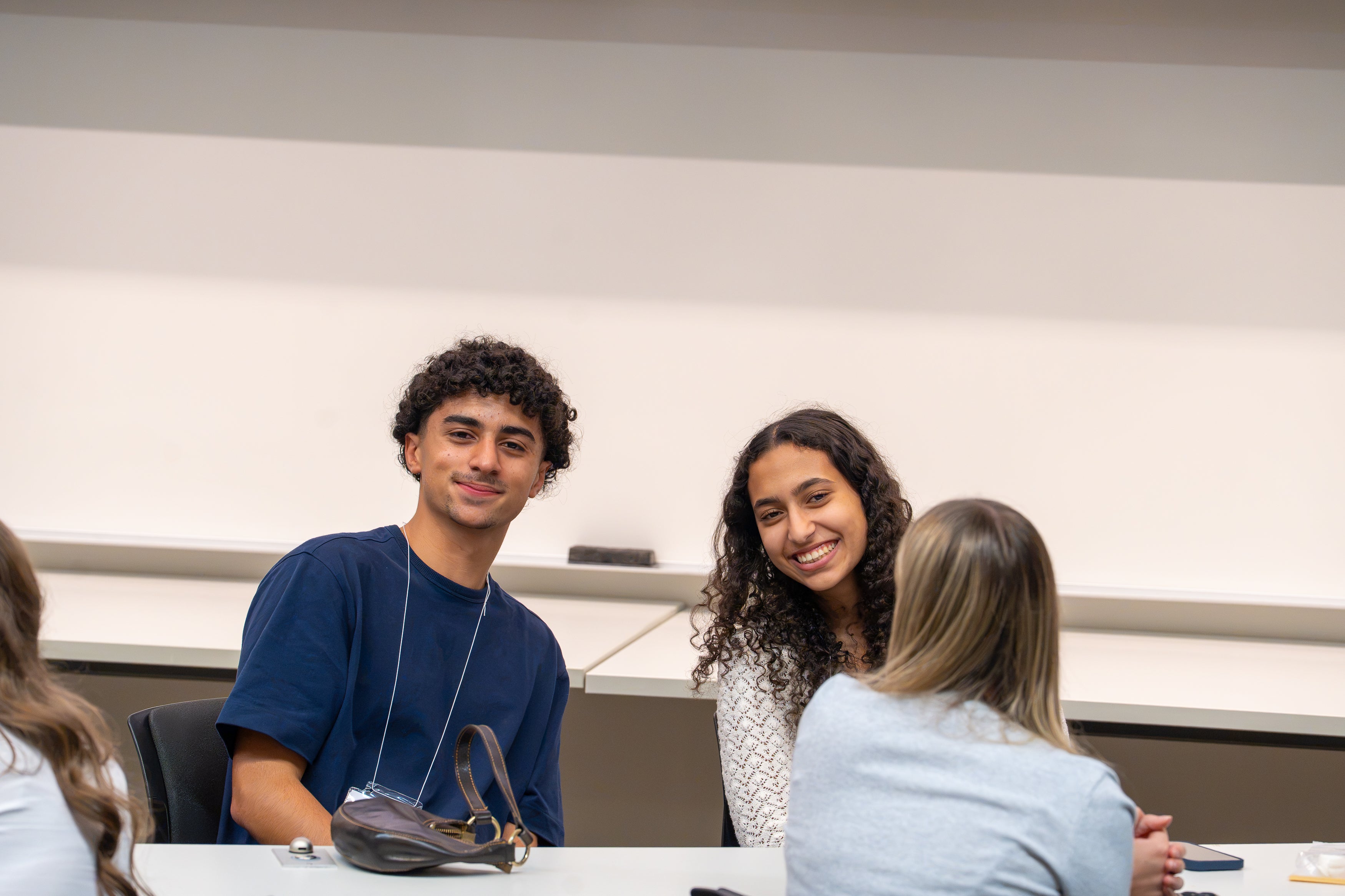 Three attendees sit together in a classroom during an event, with two facing the camera and another seated across from them at a desk.