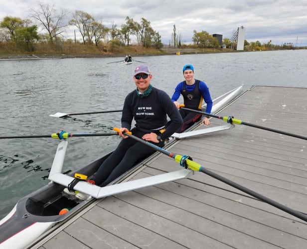 Two people sit in a rowing shell docked beside a wooden platform on a calm river under a cloudy sky. Each person holds an oar, with additional rowers visible in the distance. Trees with autumn foliage line the far bank, along with utility structures and a tall industrial-looking tower.