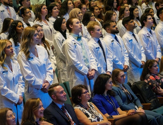 A group of students standing in an auditorium in their white coats