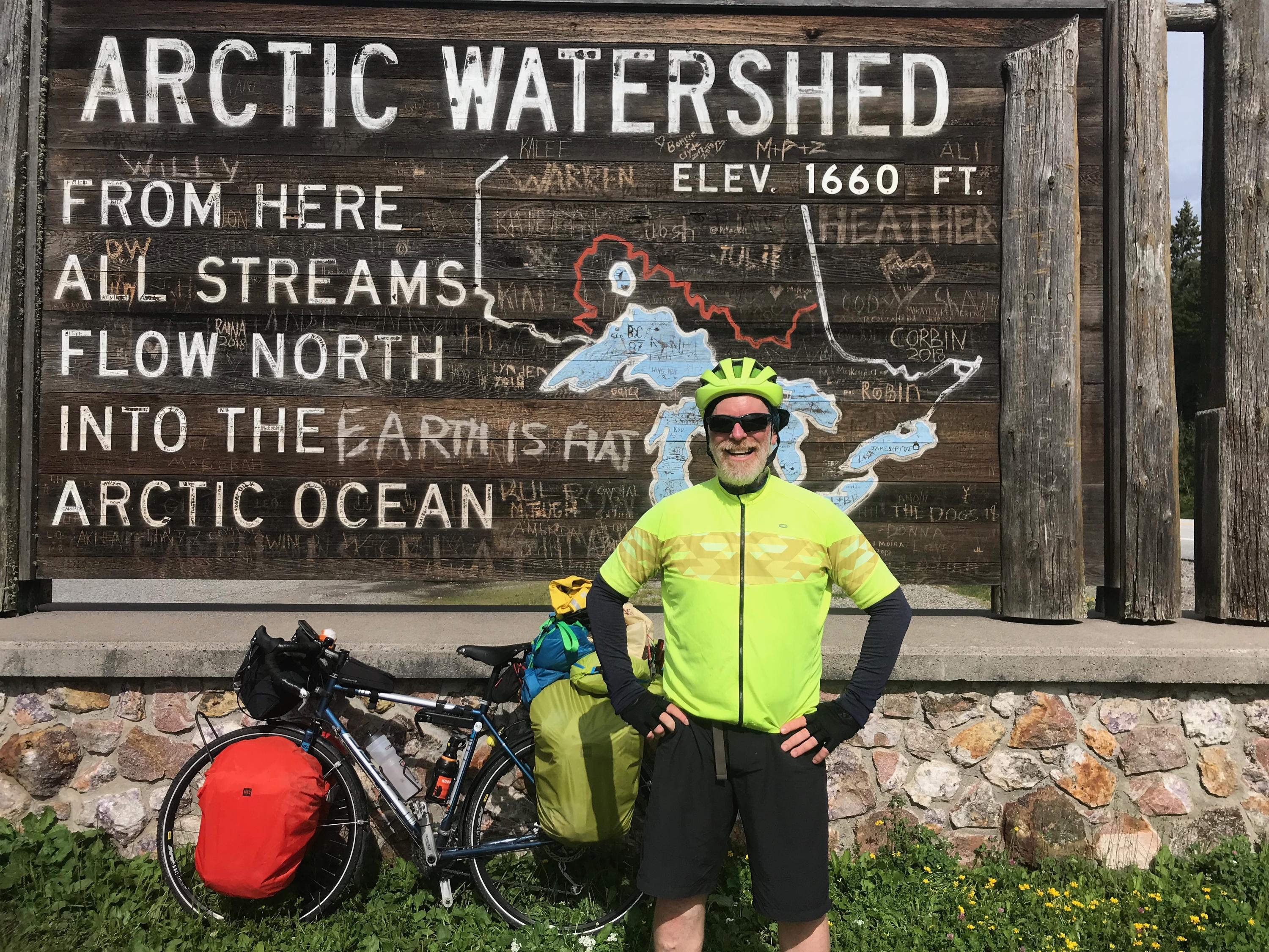 Greg Andres in front of a sign reading Arctic Watershed with a bike