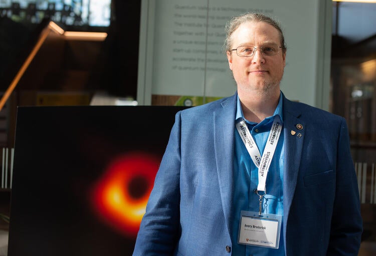 Professor Avery Broderick standing in front of a poster of the image of black hole M87*
