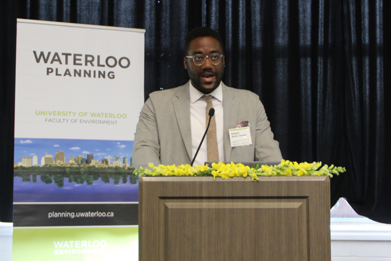 2024 Graduate Luncheon Alumni Speaker Robert Walter-Joseph presenting in front of a podium and School of Planning banner