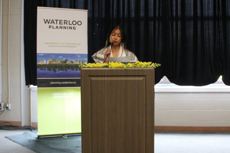 2024 Graduate Luncheon Undergradate Speaker Raida Chowdhury presenting in front of a podium and School of Planning banner