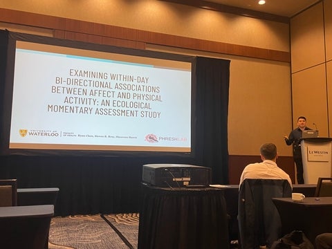 Ryan Chen standing on a podium presenting his research with the title slide displayed.