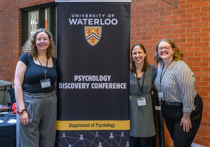 PhD students Candice Rubie and Sarah English stand besidea Psychology Discovery Conference Banner with Psychology Prof. Katherine White